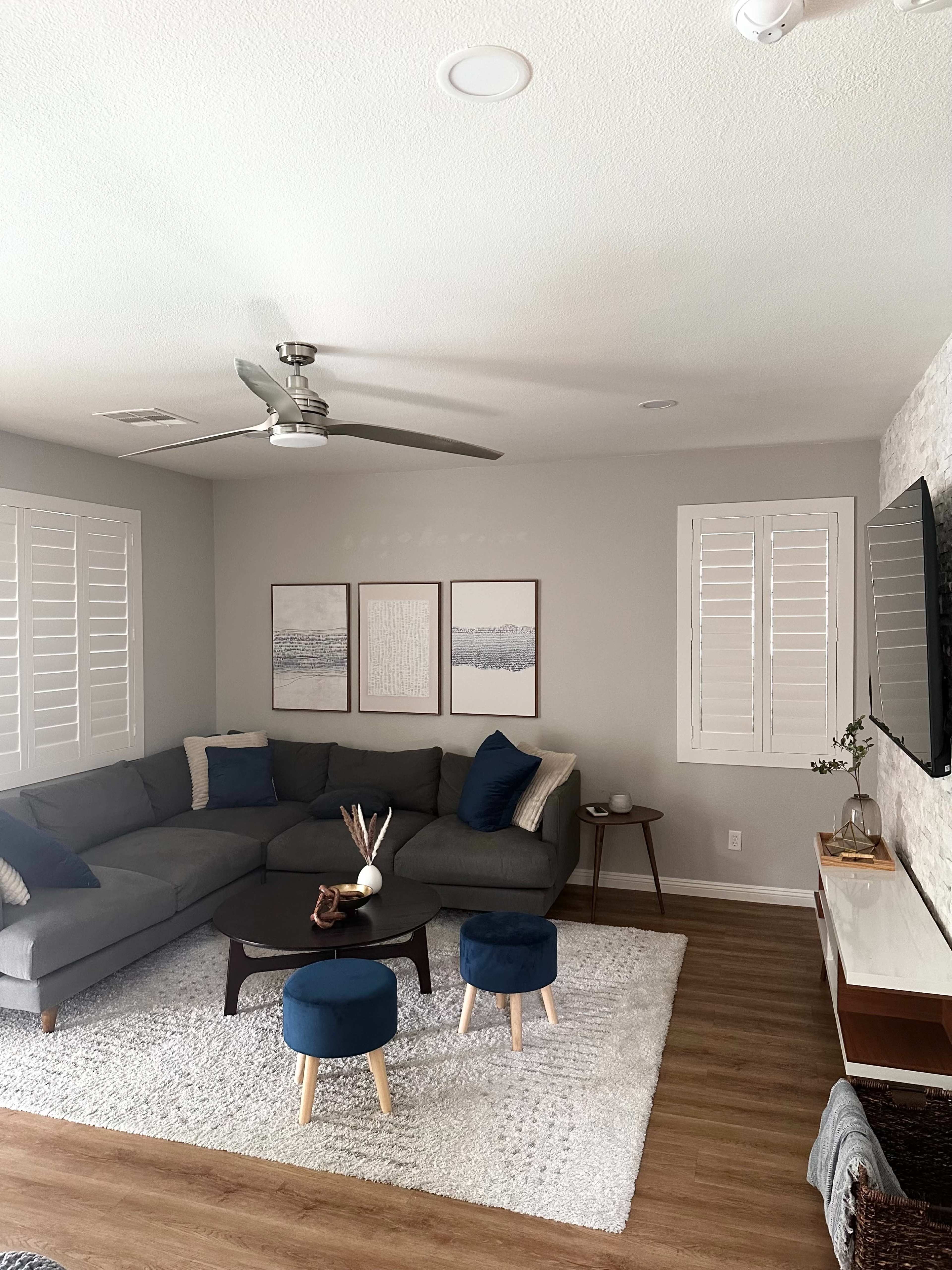 The living room features a gray sectional sofa, two blue stools, a large area rug, and three framed artworks on the wall.