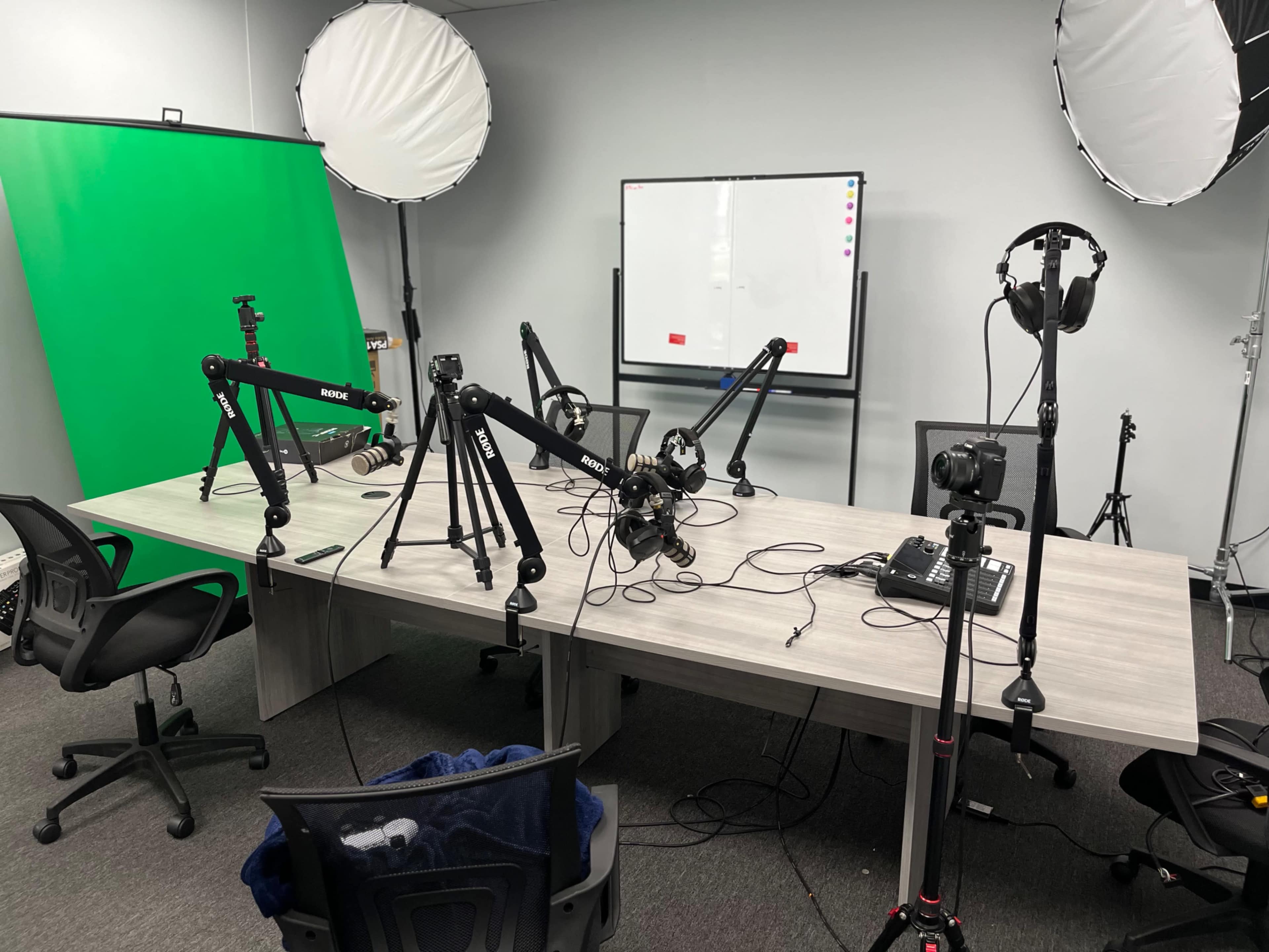 The image shows a recording studio setup with a table, several microphones on adjustable arms, lighting equipment, a green screen, and a whiteboard in the background.