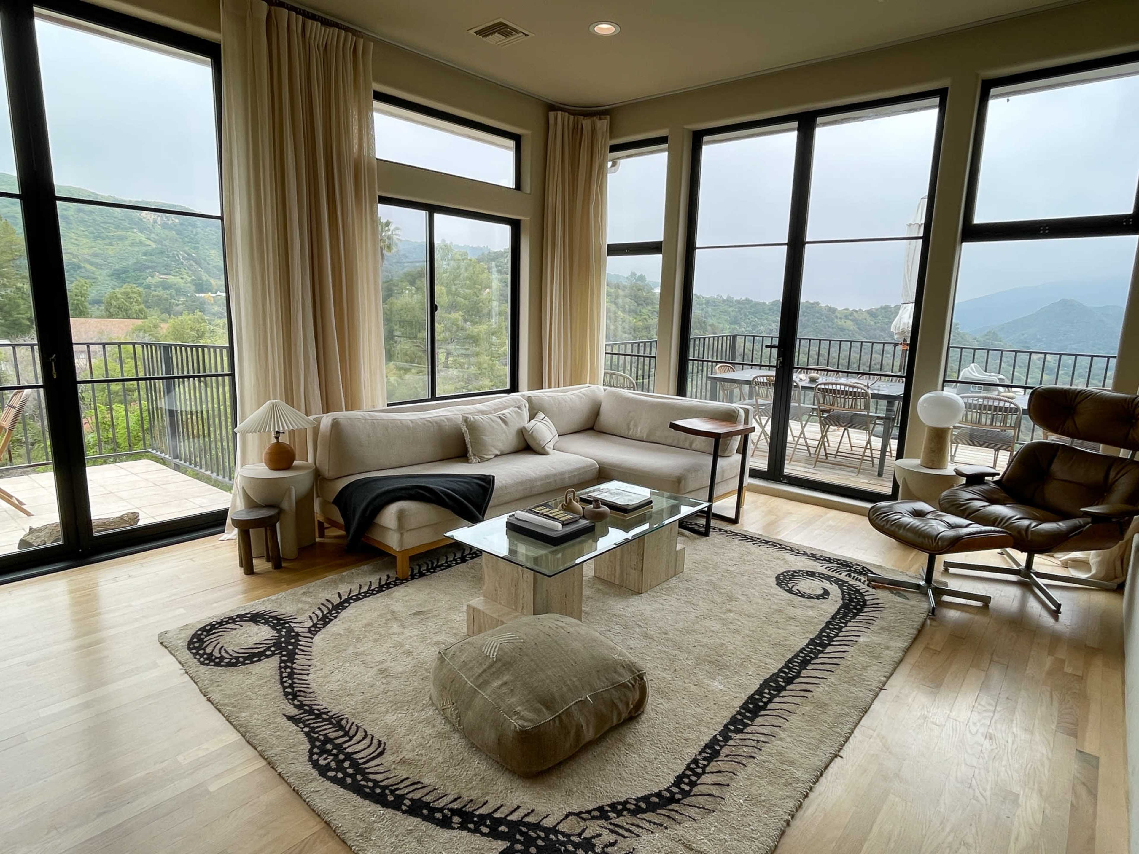 The living room features large windows with mountain views, a light-colored sofa, a glass coffee table, and a rug on the wooden floor.