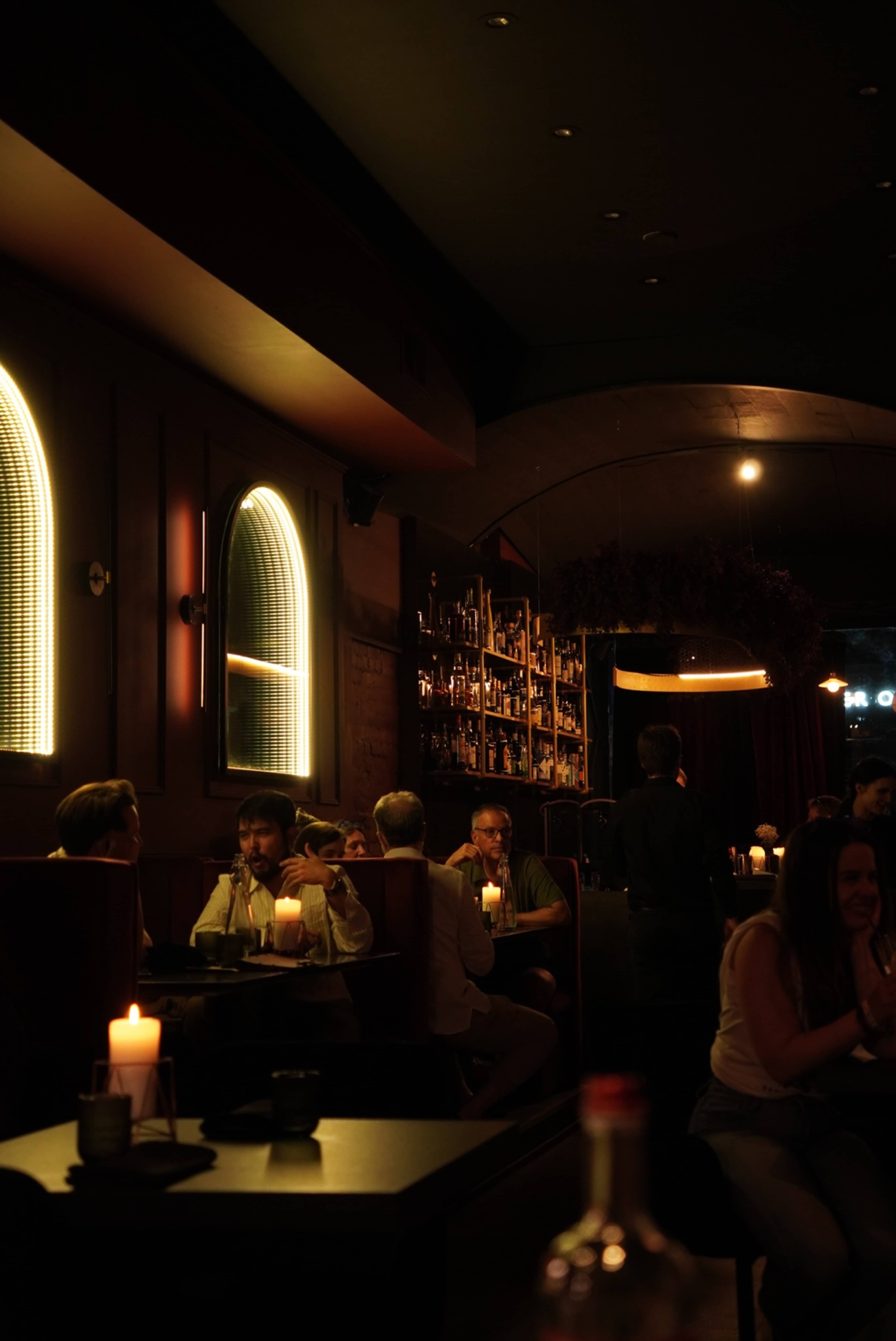 The image shows a dimly lit bar with patrons sitting at tables and a wooden bar lined with bottles in the background.