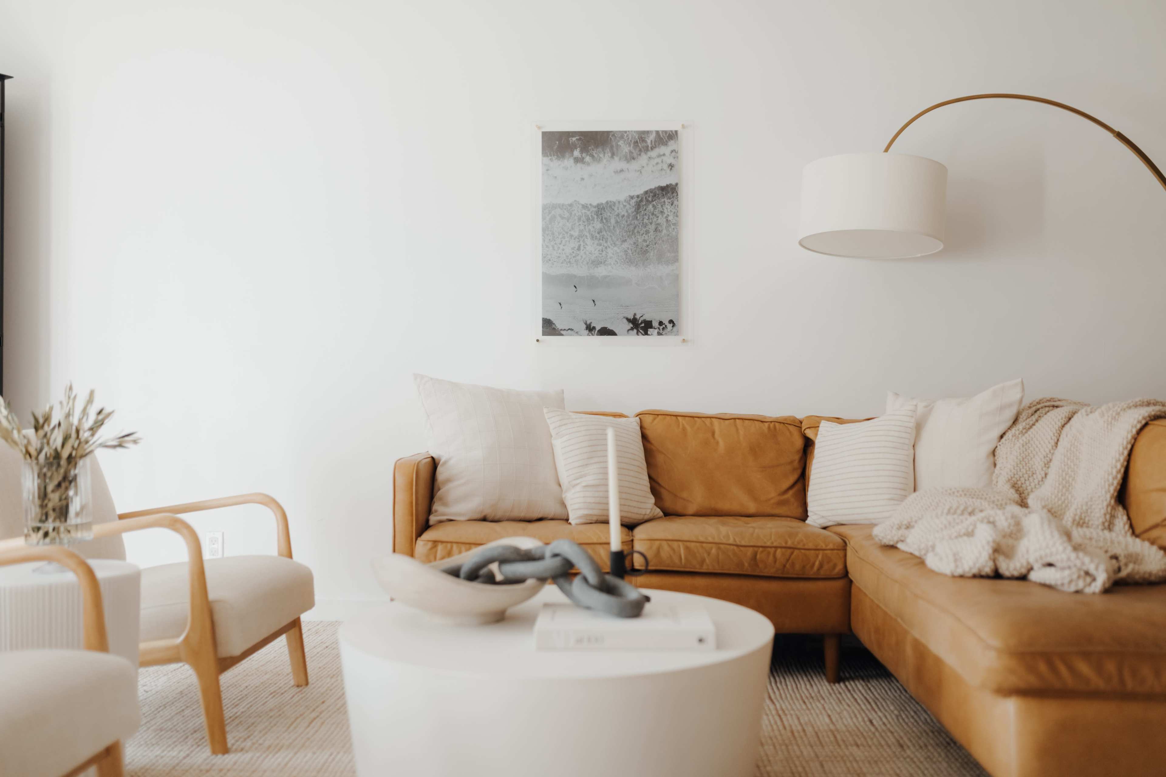 A living room features a brown leather sofa with decorative pillows, a round white coffee table, and a floor lamp, all set against a white wall adorned with a framed artwork.