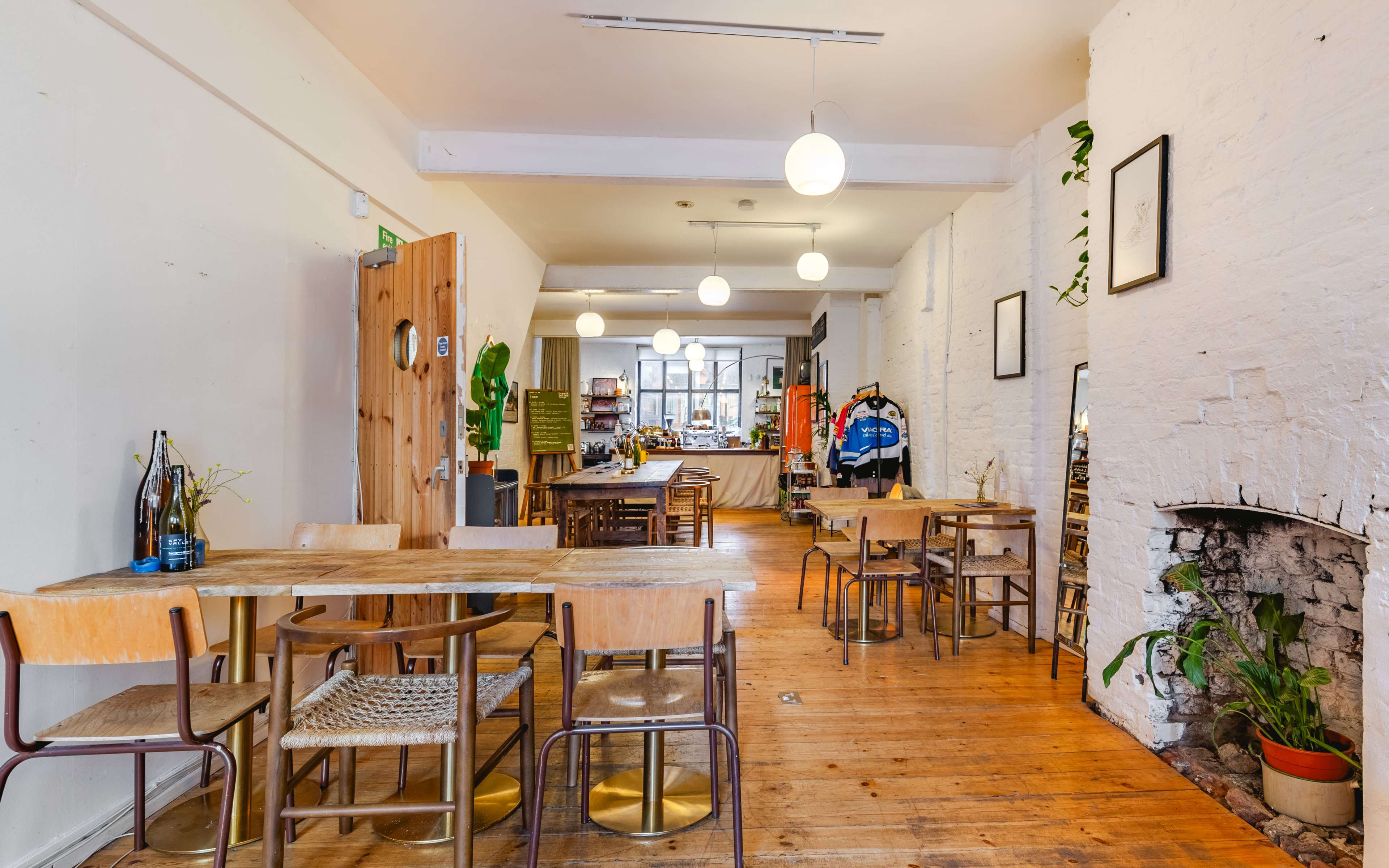 A bright, open interior of a café or dining area with wooden tables, chairs, potted plants, and a large window at the far end.