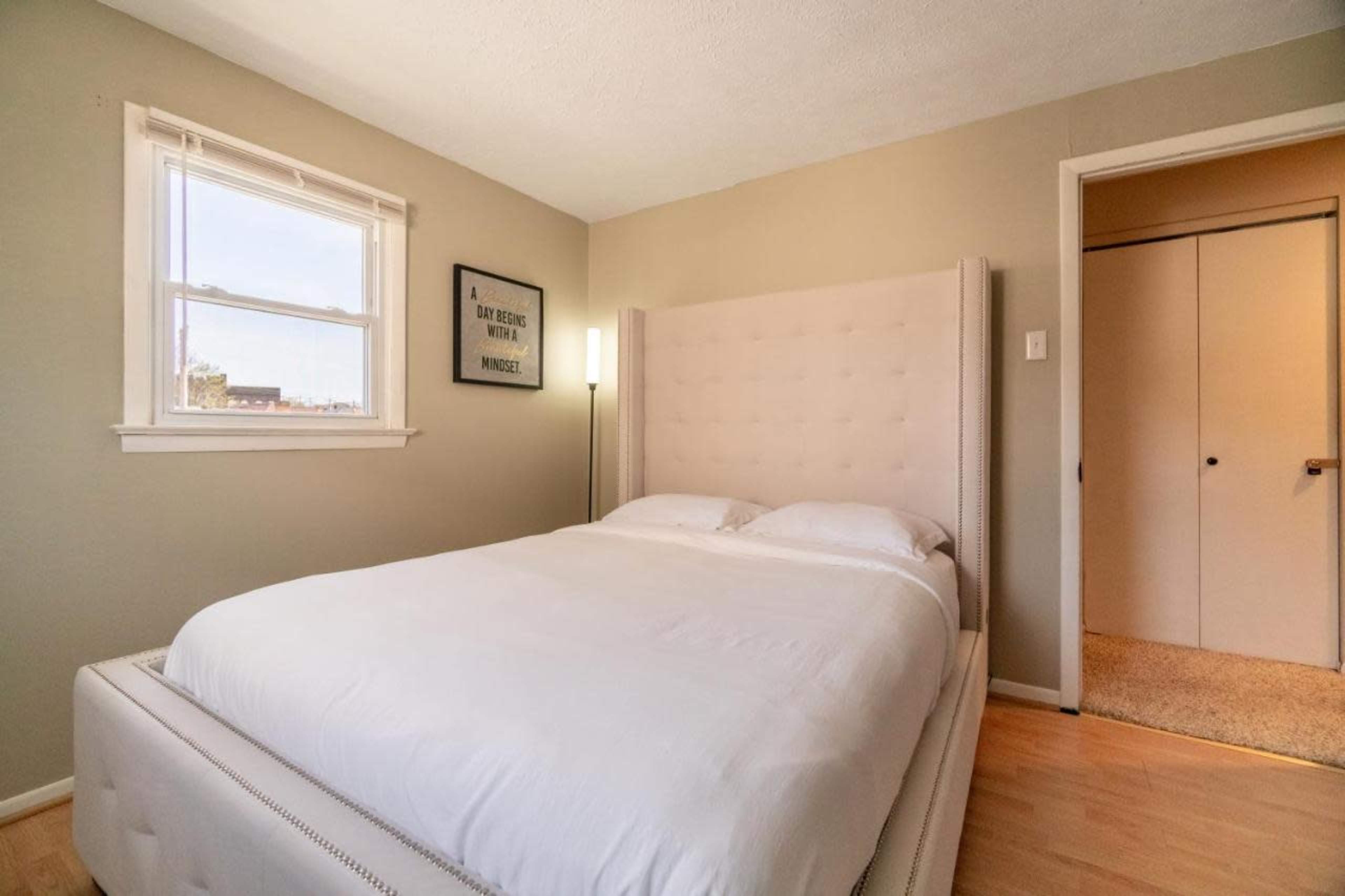 The image shows a neatly made bed in a small bedroom with light-colored walls and a window.