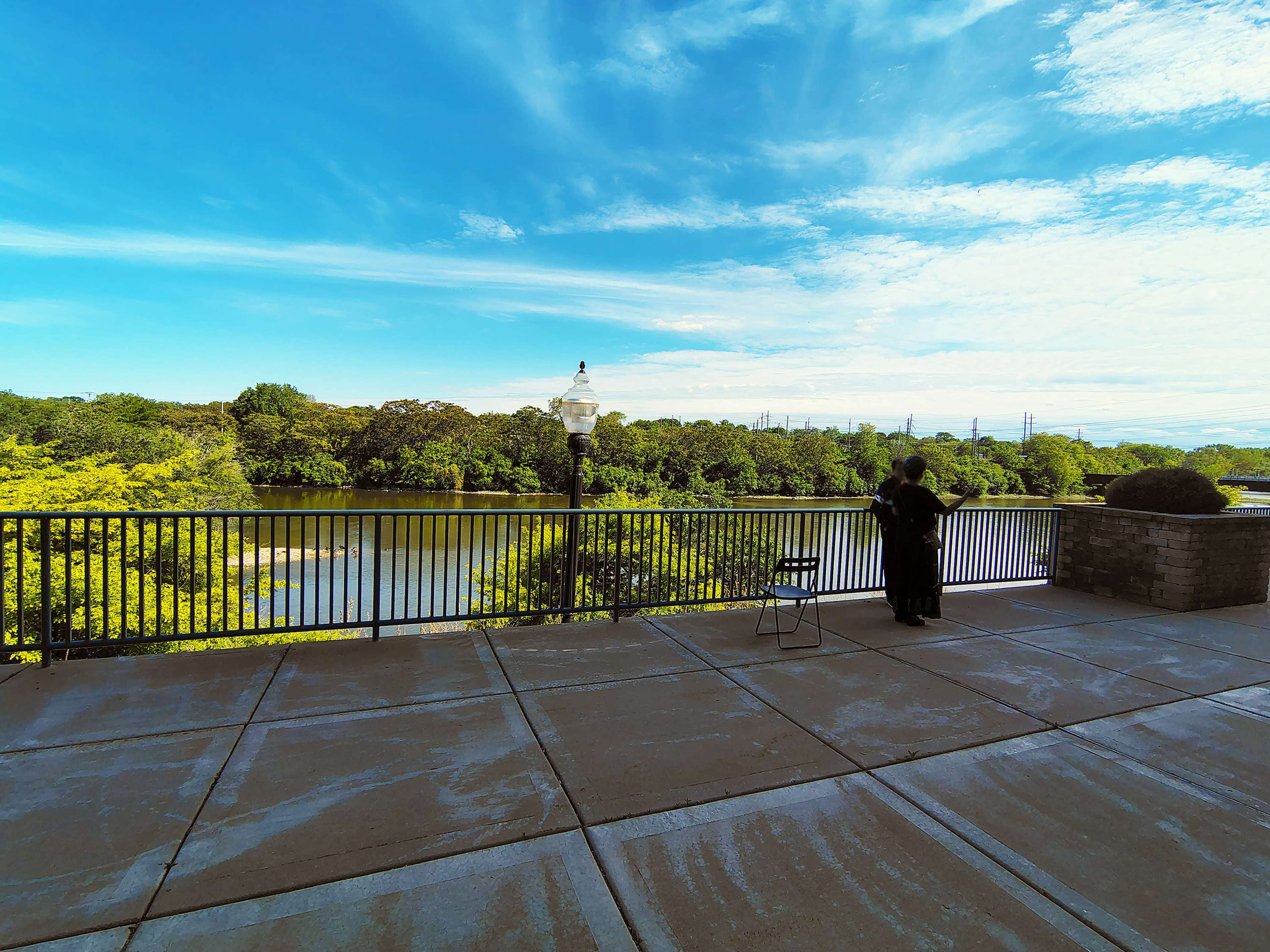 A person stands by a railing overlooking a river, surrounded by trees and a clear blue sky.