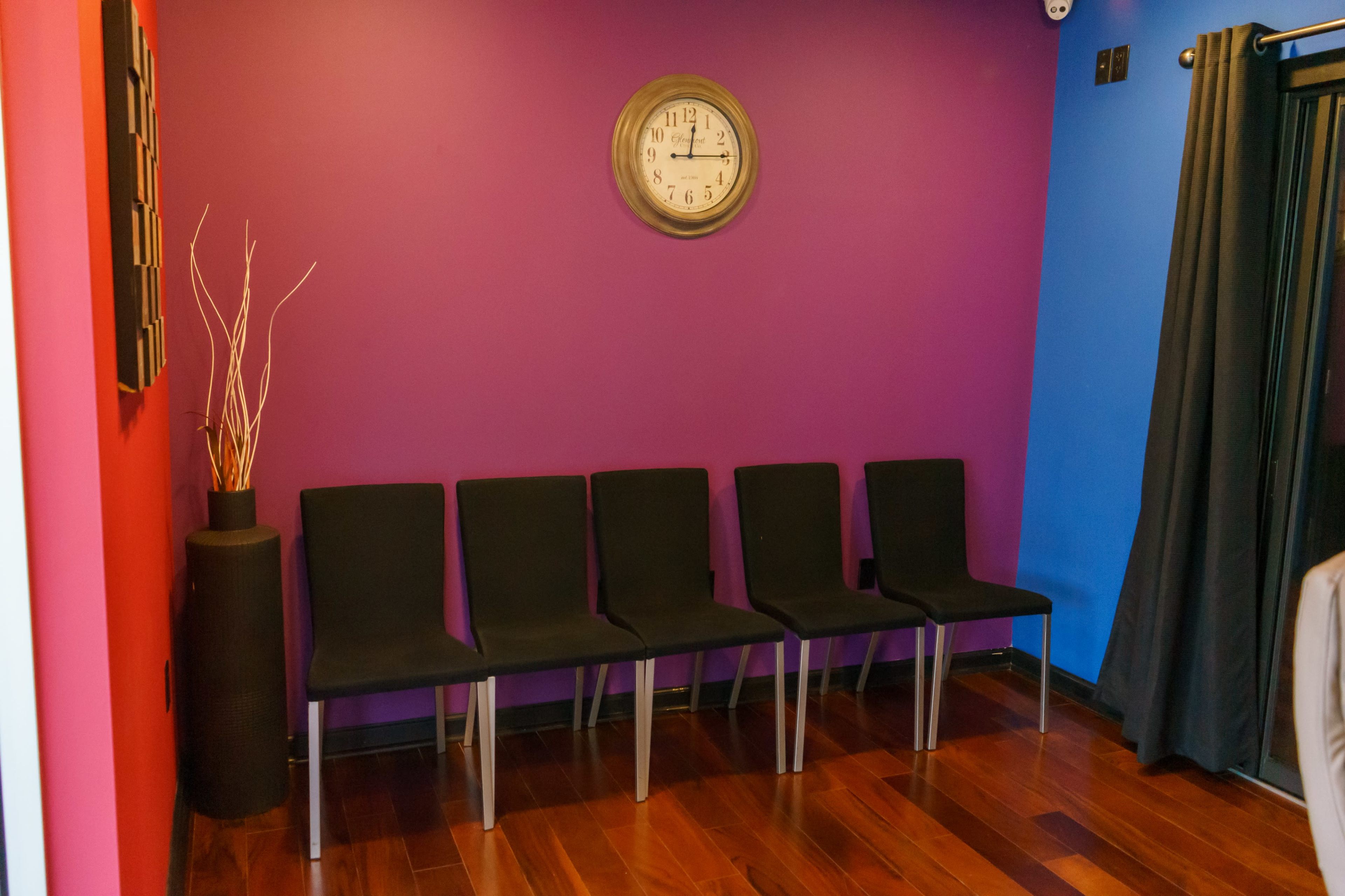 A waiting area with five black chairs against a purple wall, a clock on the wall, and a decorative plant in a black pot.