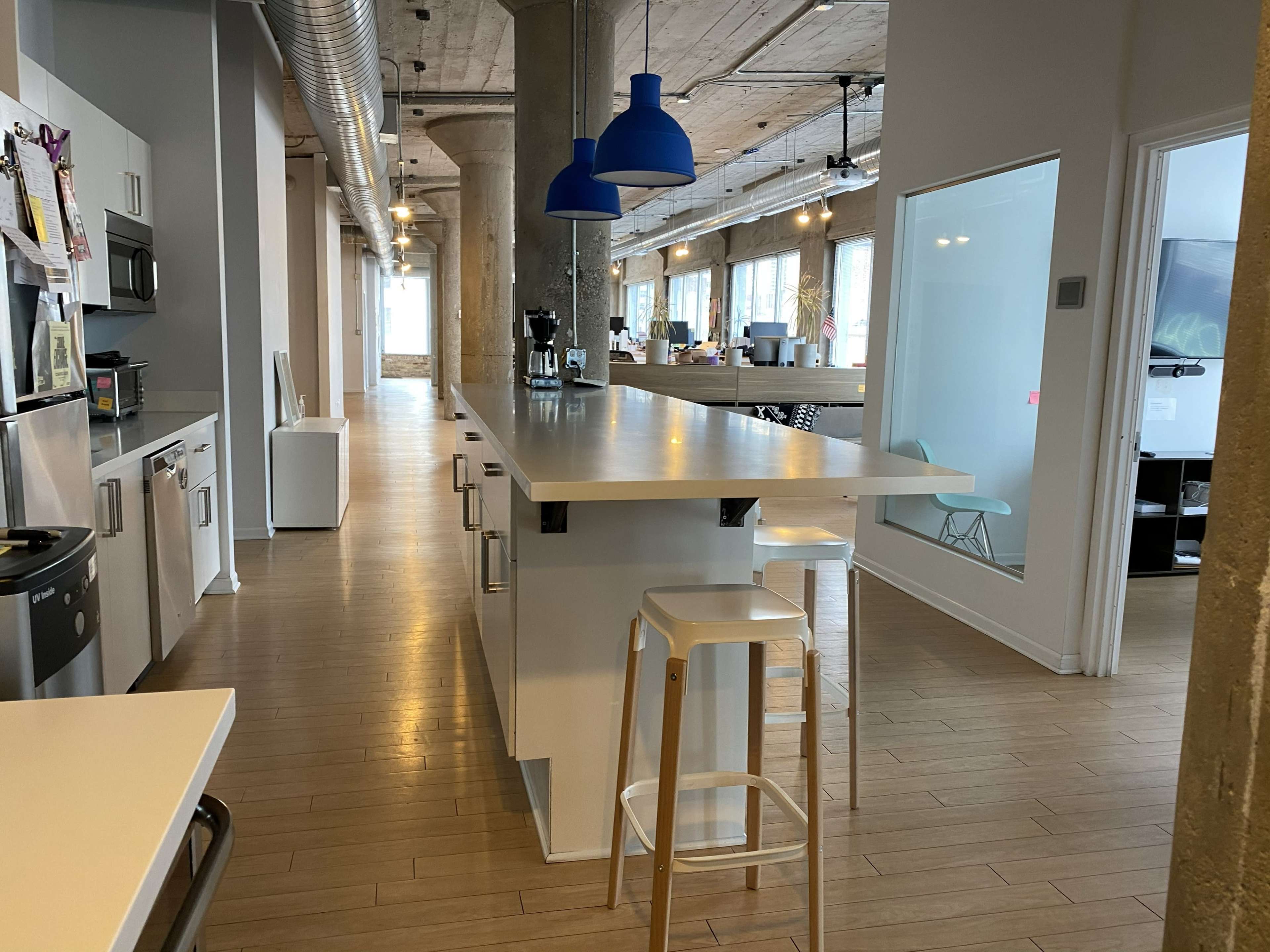 The image shows a modern, open office space with a kitchen area featuring a long white countertop, stainless steel appliances, and blue pendant lights.