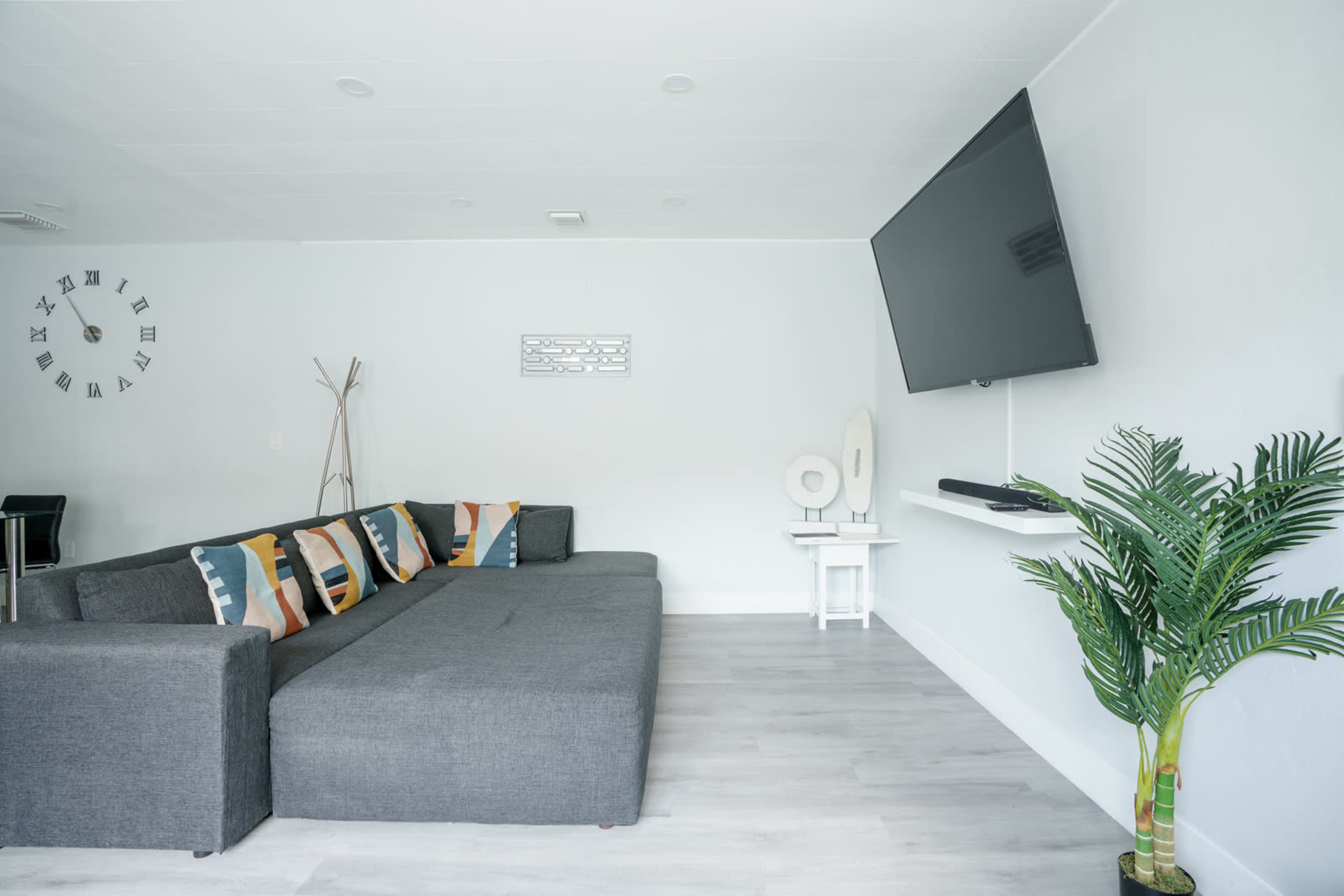 The living room features a gray sectional sofa, a mounted television, a small white side table, and a potted plant in the corner.