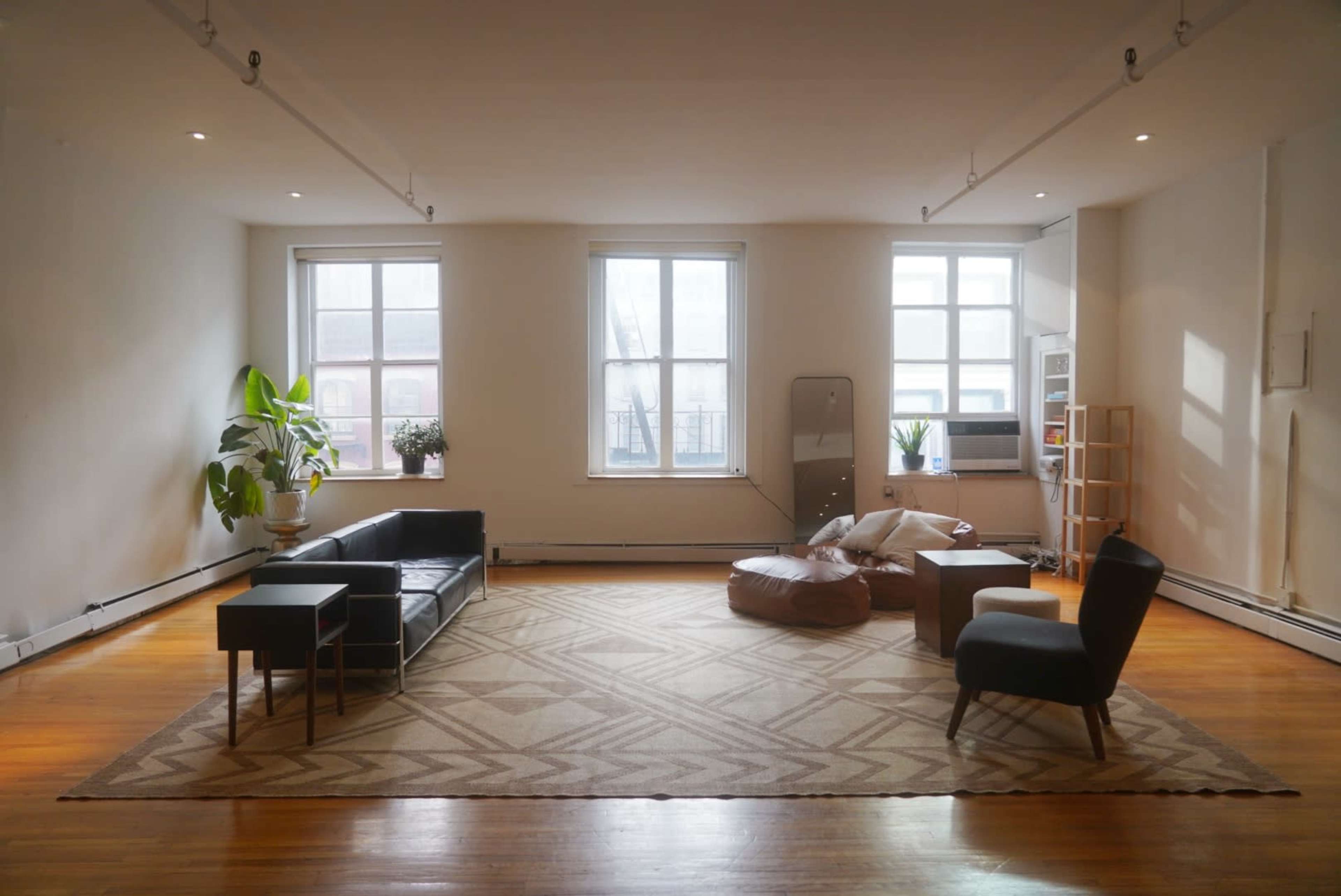 A spacious living room with a dark couch, a coffee table, a beanbag chair, and large windows letting in natural light.