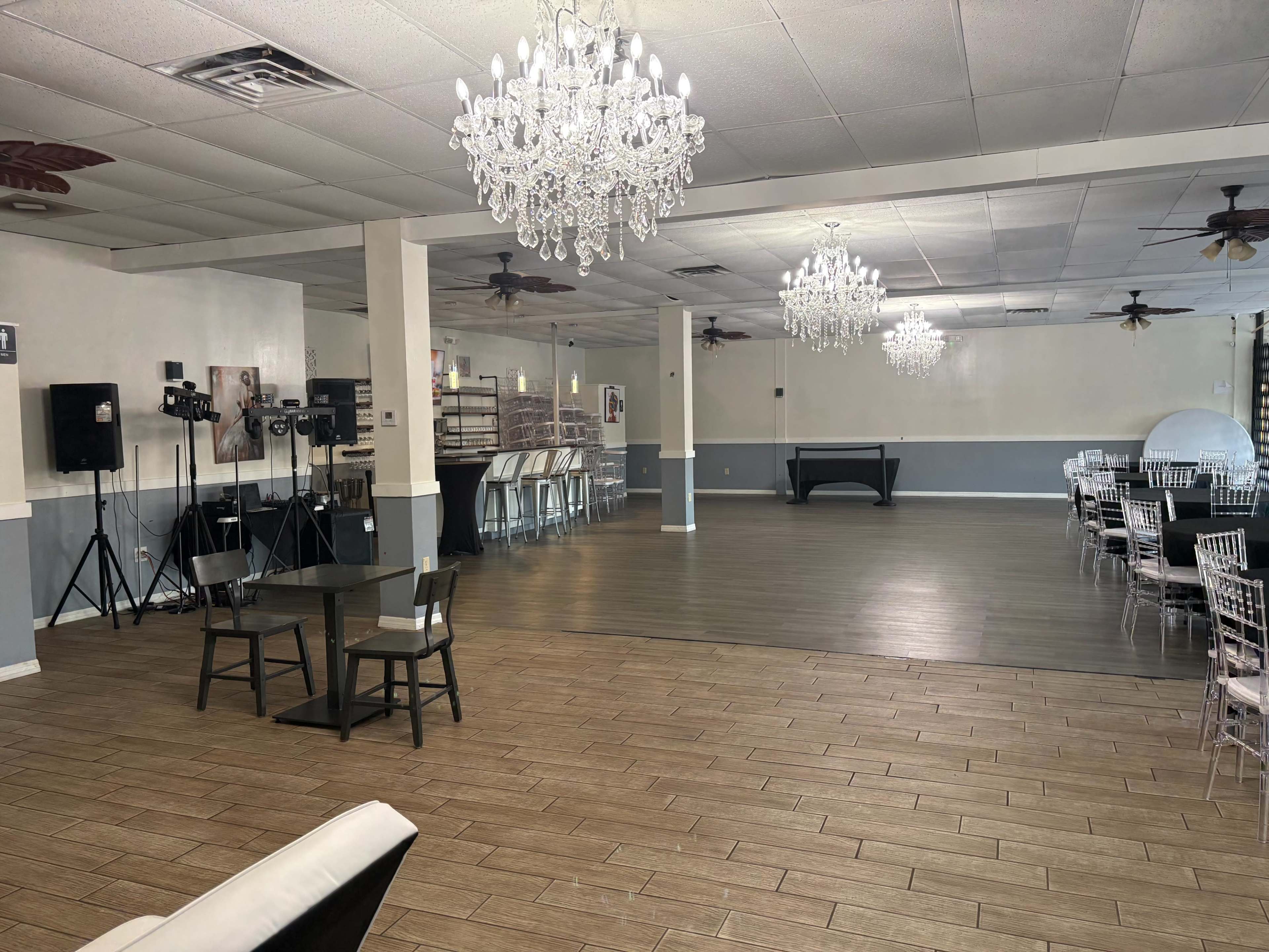 The image shows a spacious event hall featuring multiple chandeliers, wooden flooring, and a seating area with tables and chairs arranged for an event.