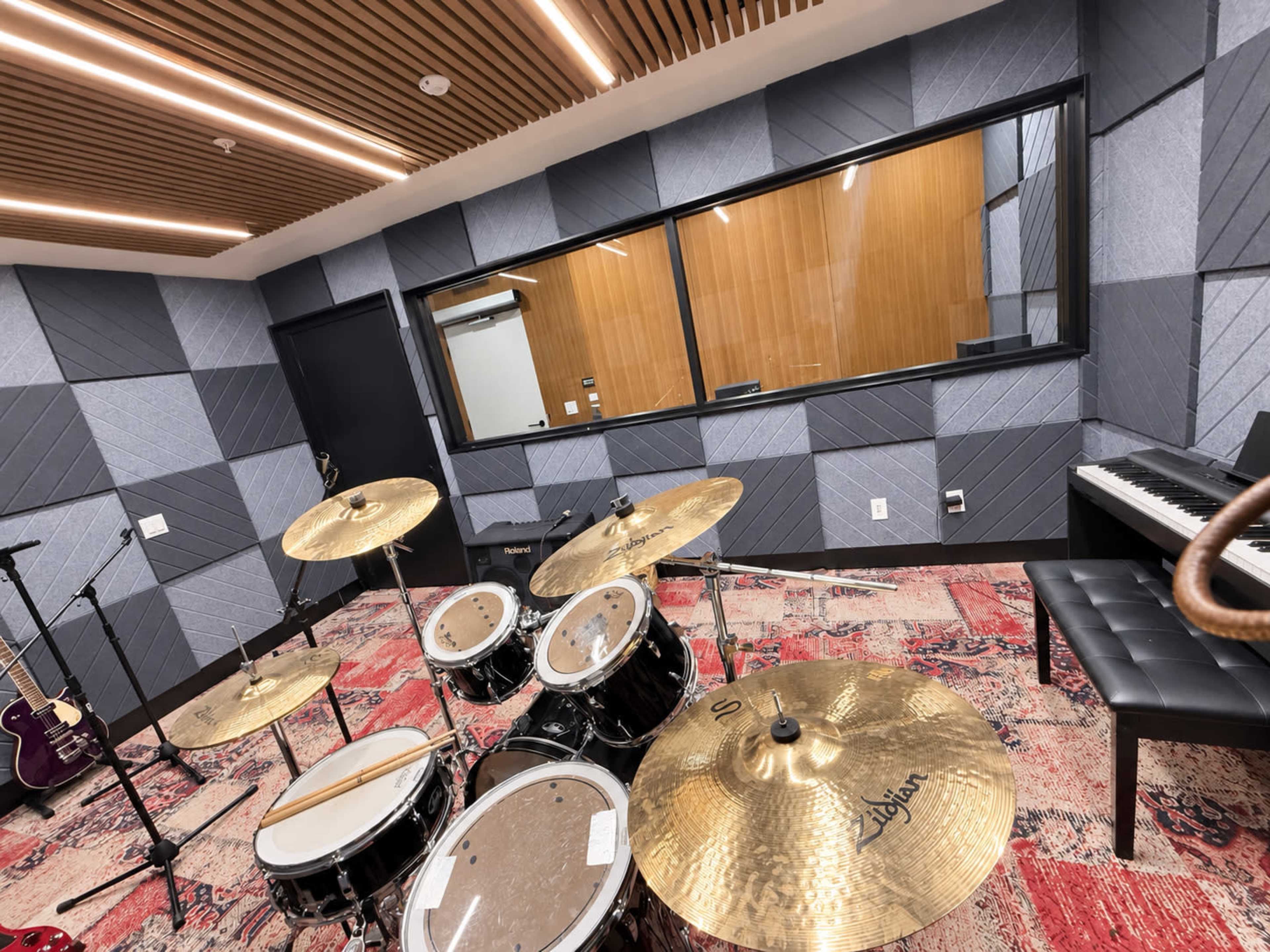 The image shows a music practice room featuring a drum set, a piano, and soundproof glass windows.