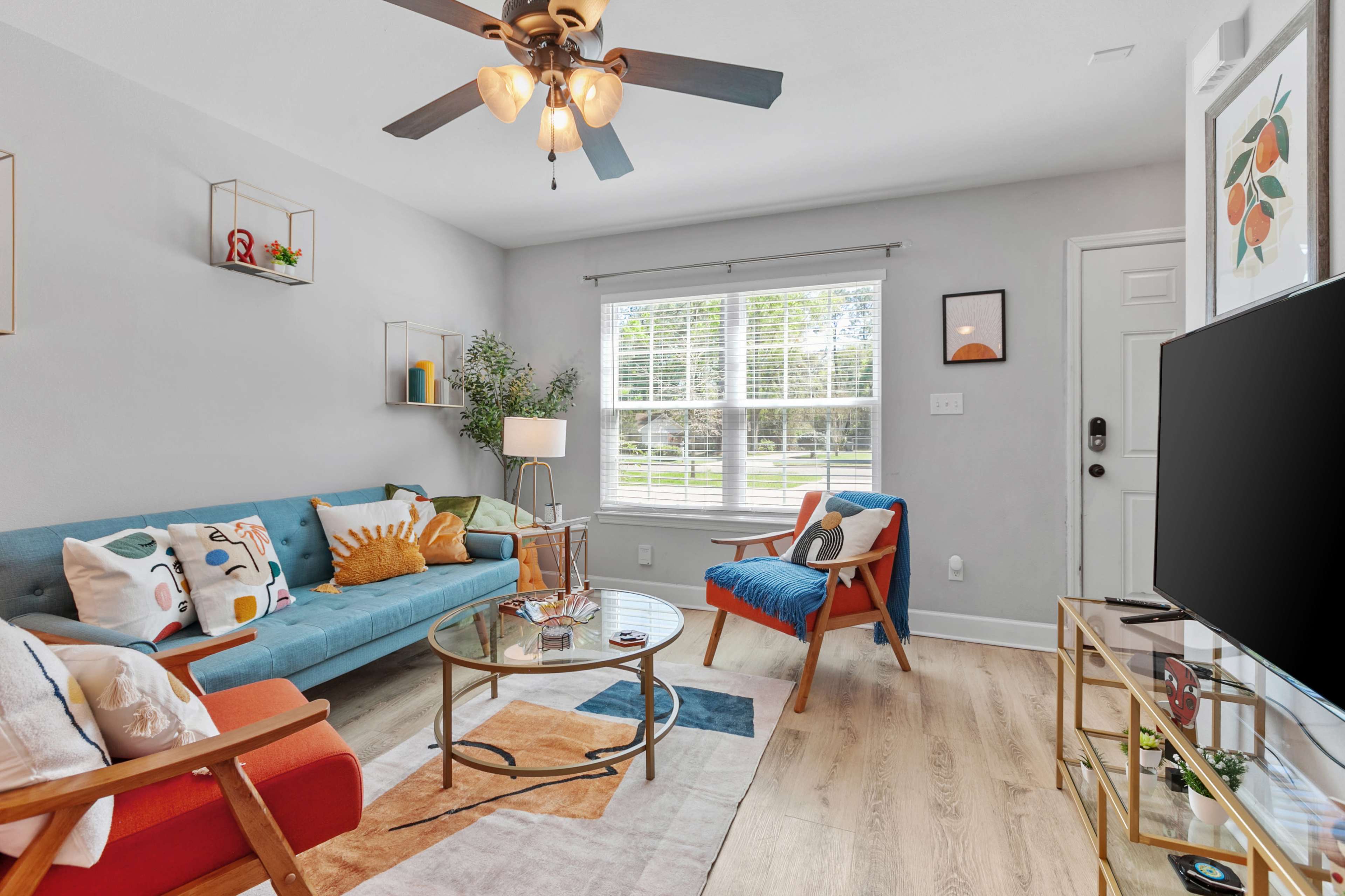A modern living room features a blue sofa, an orange armchair, a glass coffee table, and large windows allowing natural light to enter.
