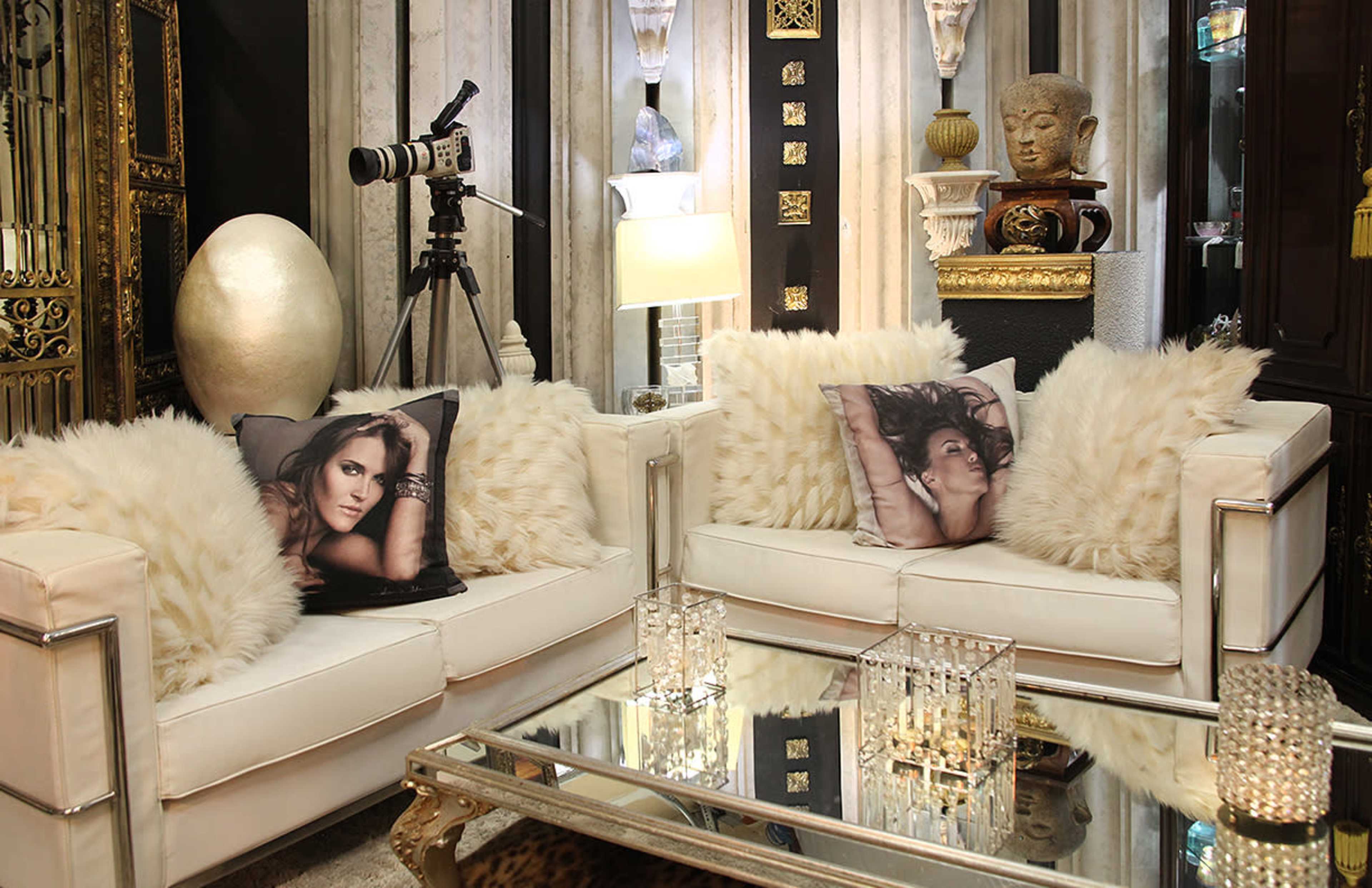 The image displays a stylish living room featuring two white sofas adorned with fluffy pillows, a glass coffee table, and decorative elements such as a camera tripod and an ornate statue.