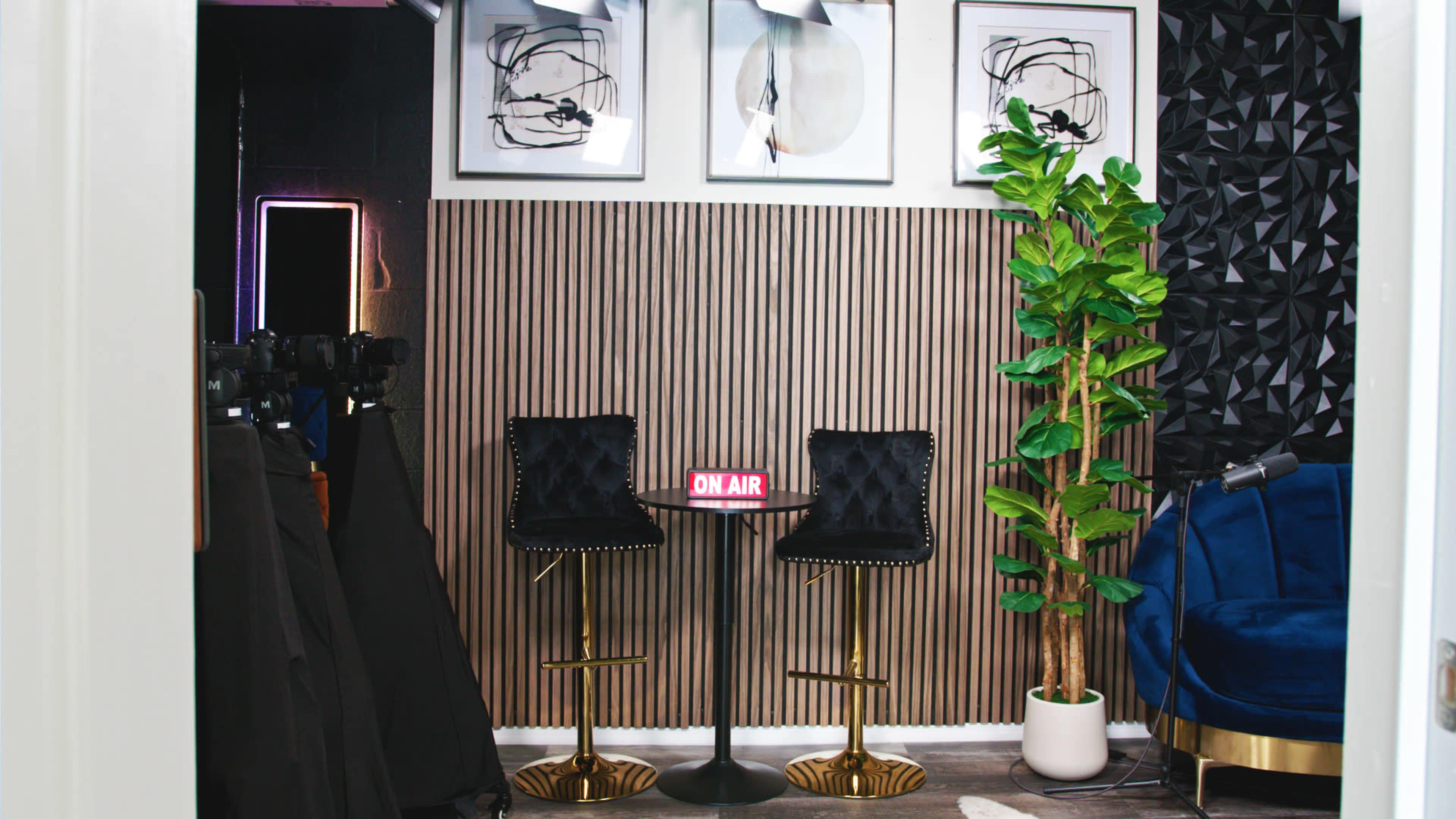 The image shows a modern studio setup with a small round table and two black upholstered stools, framed art on the wall, and a potted plant, all surrounded by a dark backdrop.