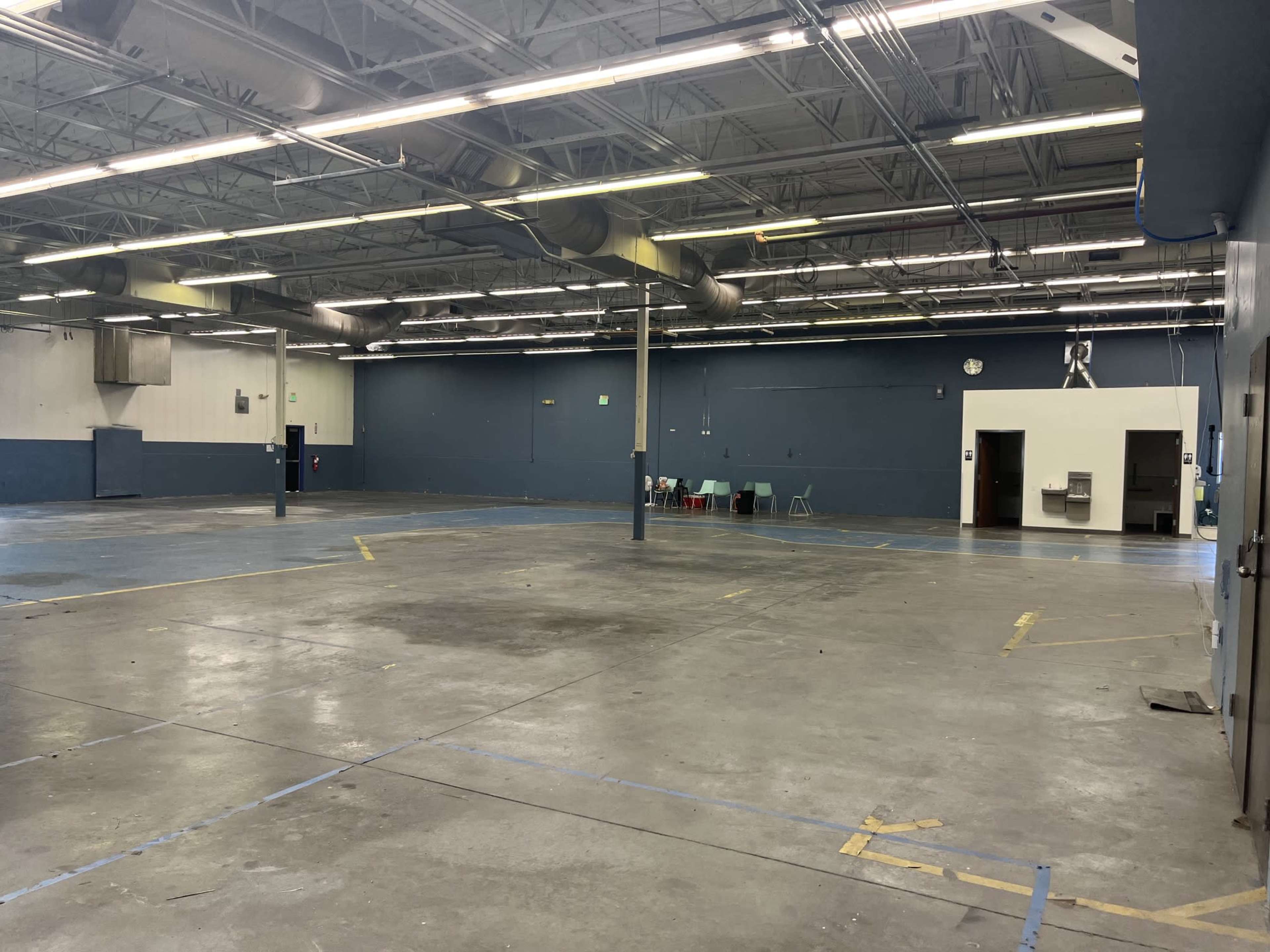 The image shows a large, empty industrial space with concrete flooring, blue walls, and visible overhead ventilation ducts.