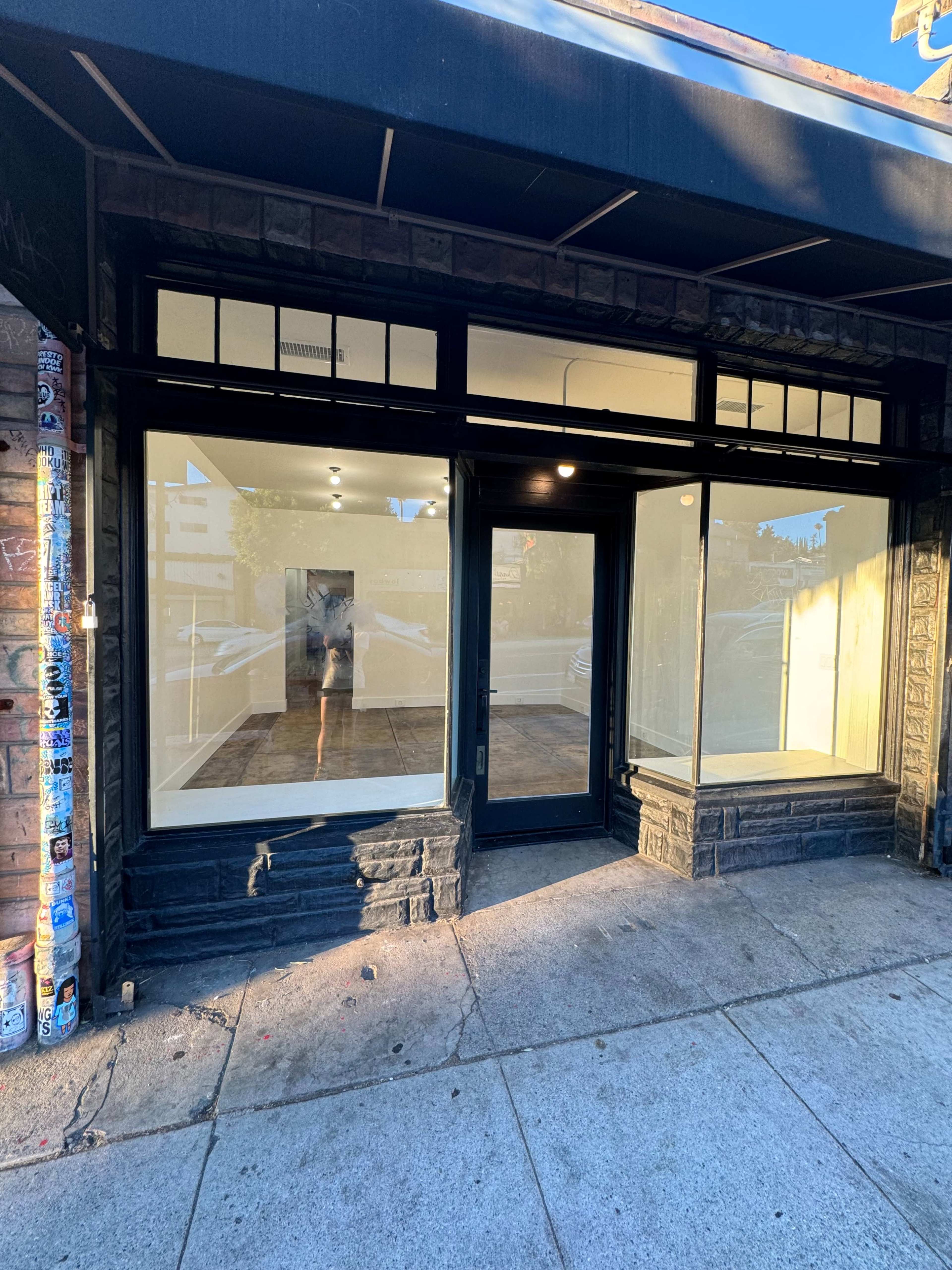 The image shows a vacant storefront with large windows and no visible signage, situated on a sidewalk.