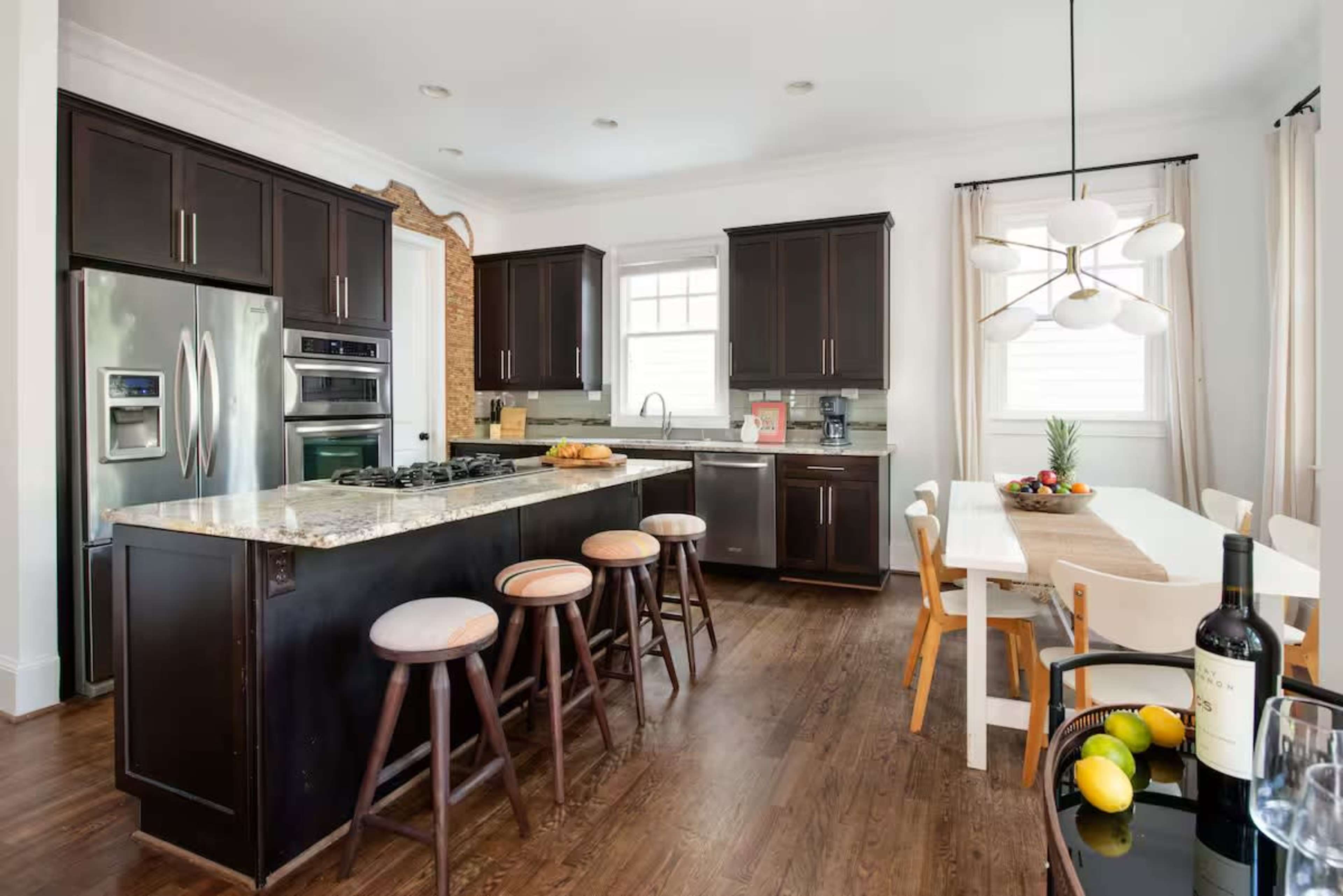 The image shows a modern kitchen with dark cabinets, a large island with bar stools, and a dining area featuring a long white table.