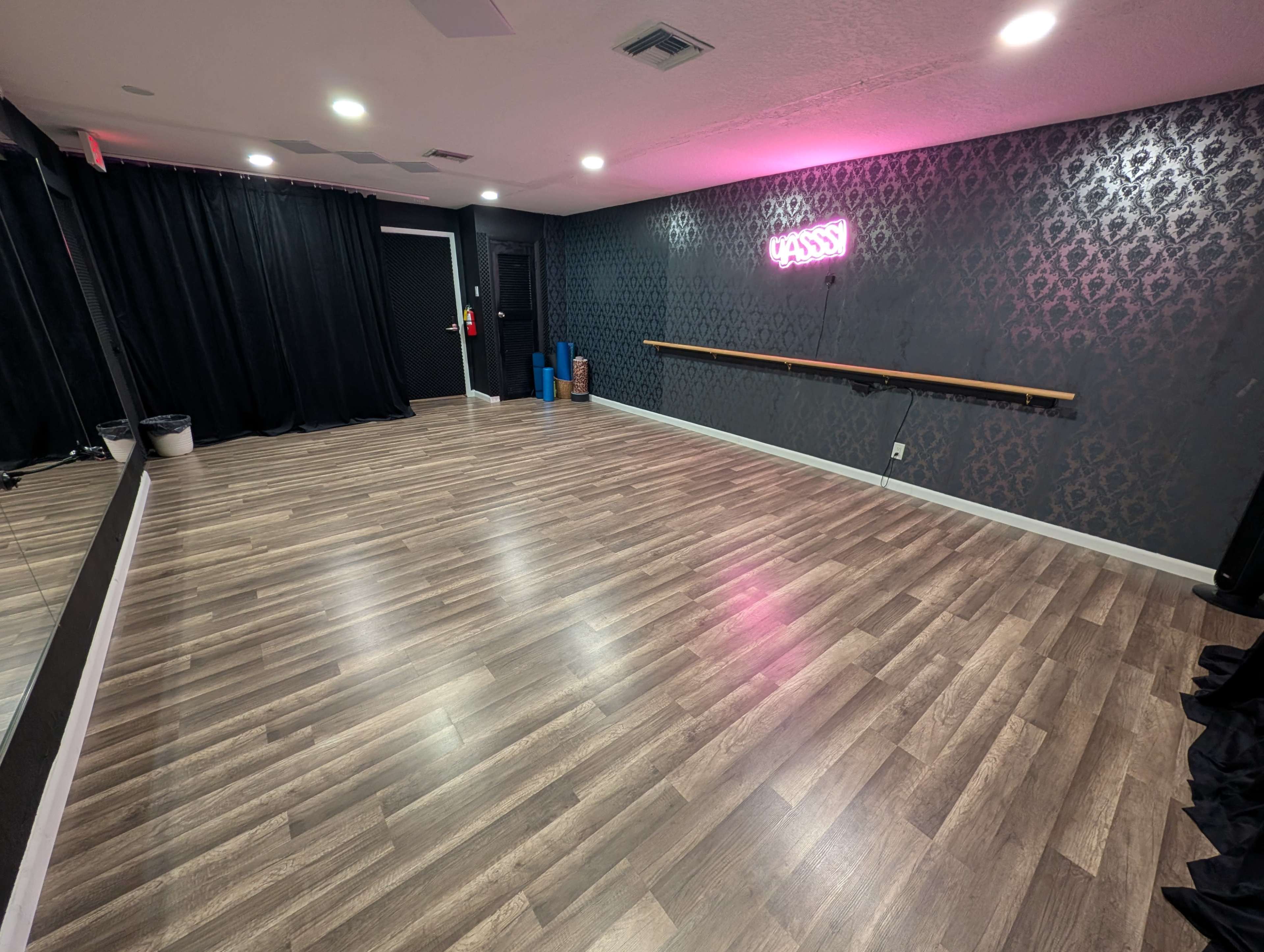 The image shows a spacious dance studio featuring wooden flooring, a mirrored wall, and pink neon signage that reads "CLASSY."