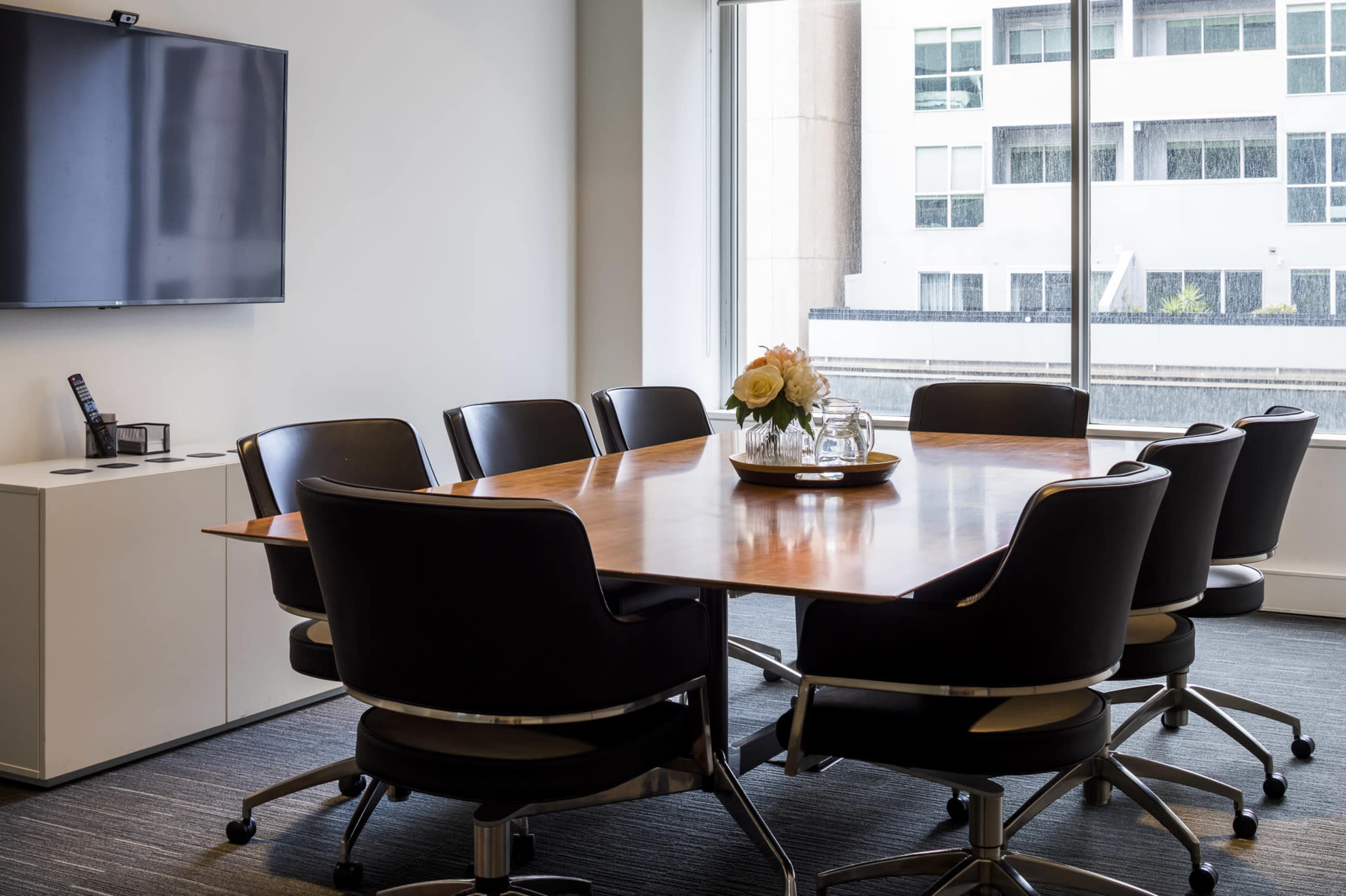 A modern conference room features a wooden table surrounded by eight black chairs, with a television mounted on the wall and large windows displaying an urban view.