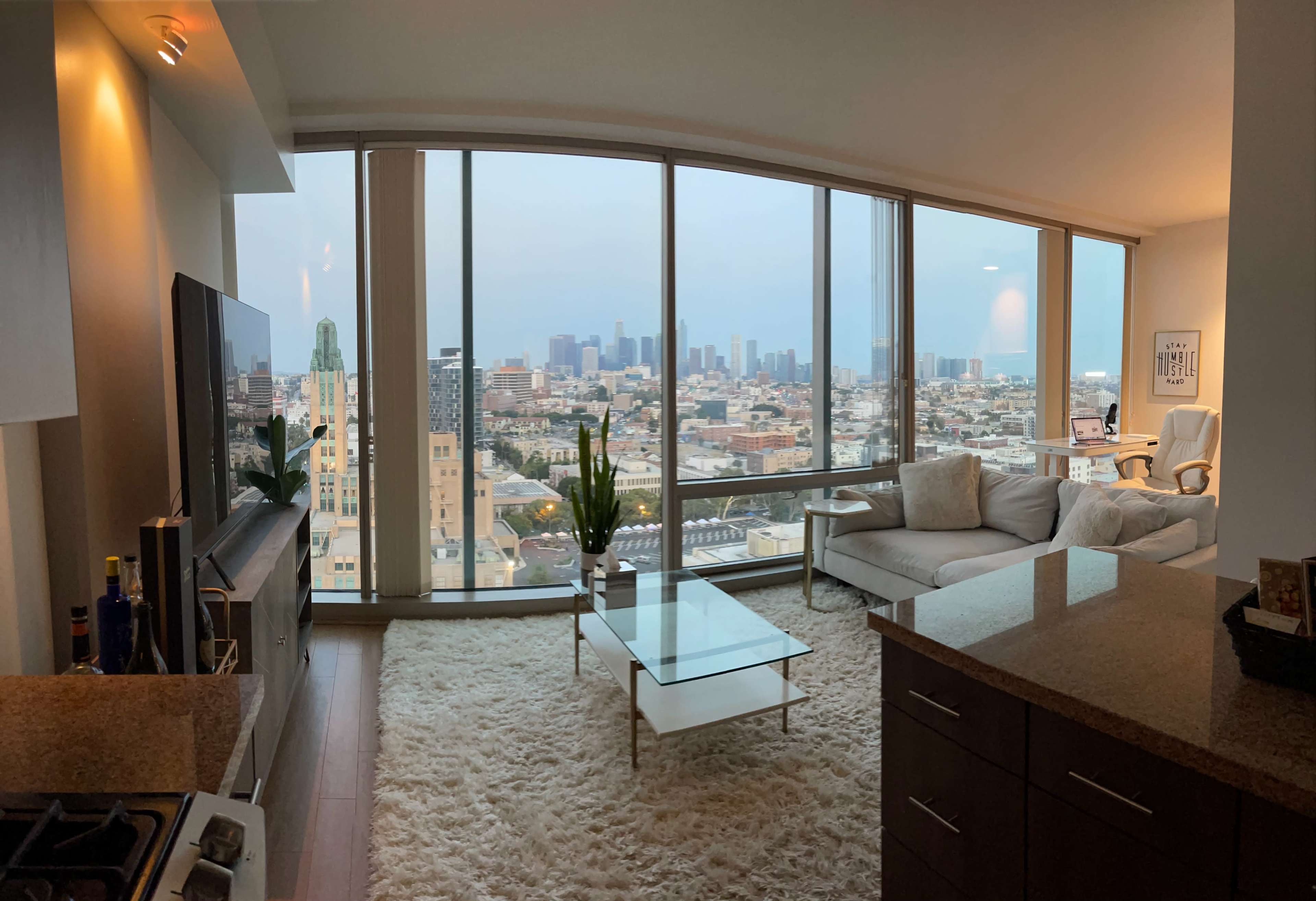 A modern living room with large windows overlooking a city skyline, featuring a couch, a glass coffee table, and a small potted plant.