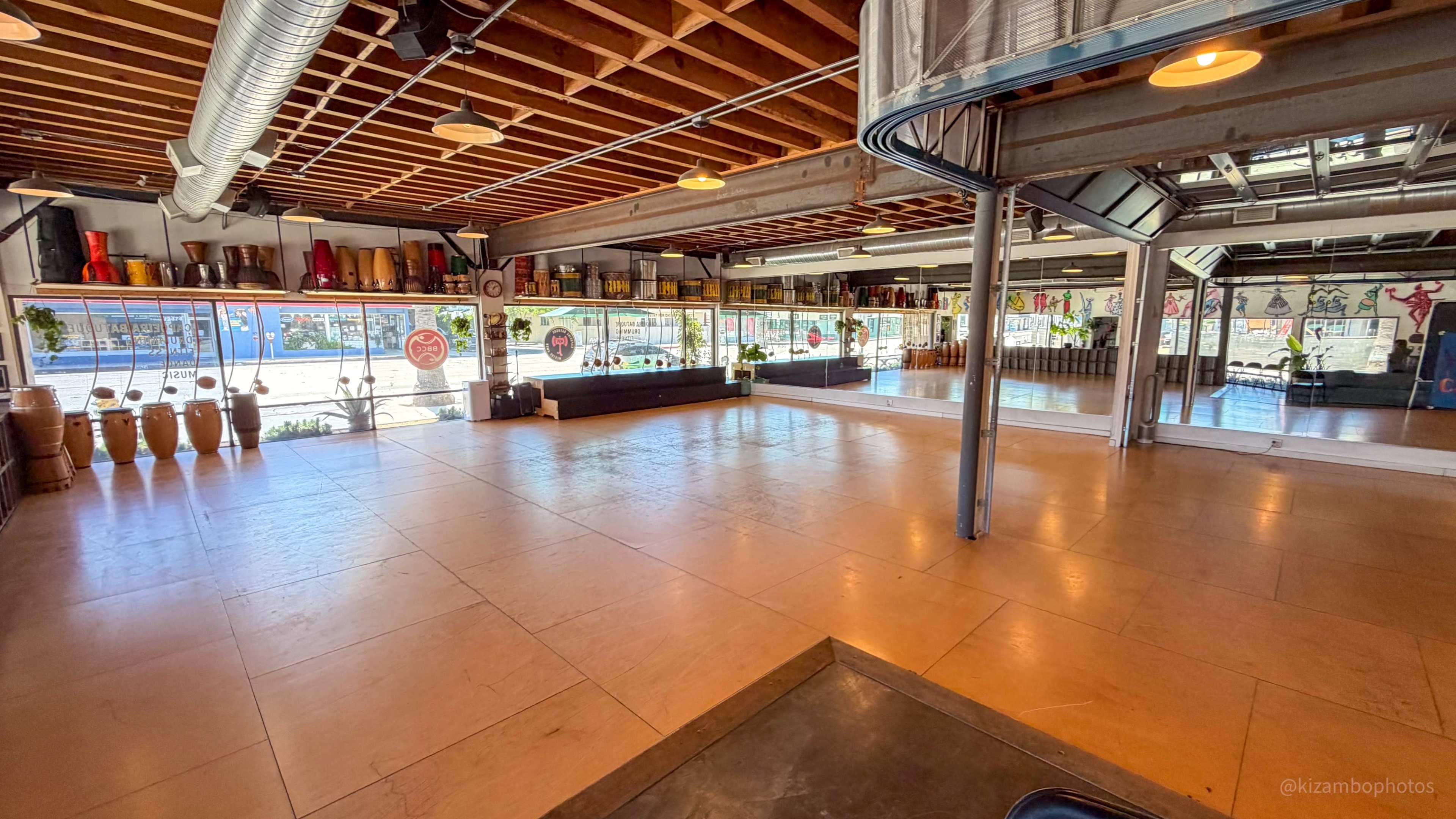The image shows a spacious interior with polished floors, large windows, and various decorations along the walls, including colorful items and drums.