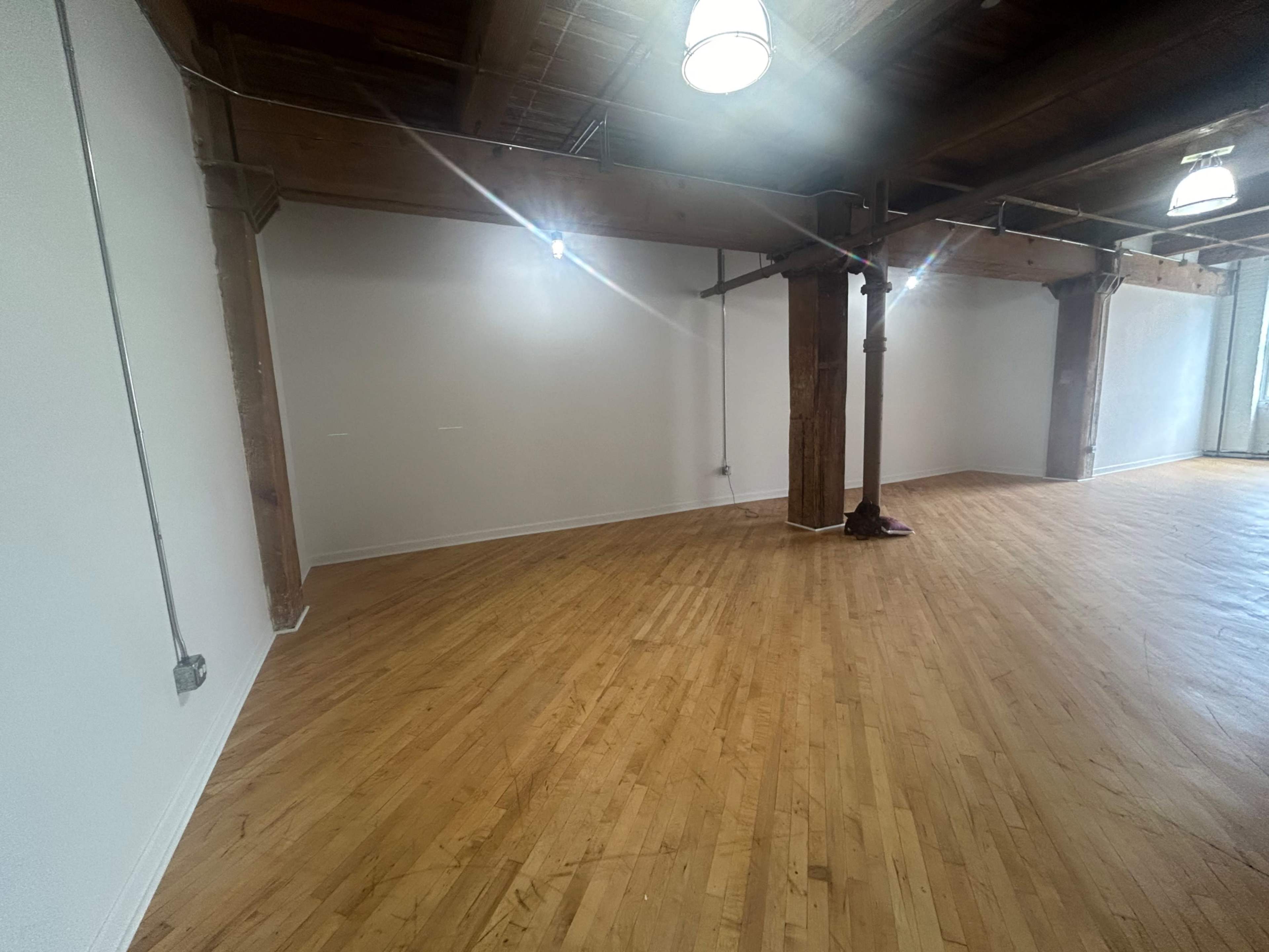 An empty room with wooden floors, exposed beams, and bright overhead lights.