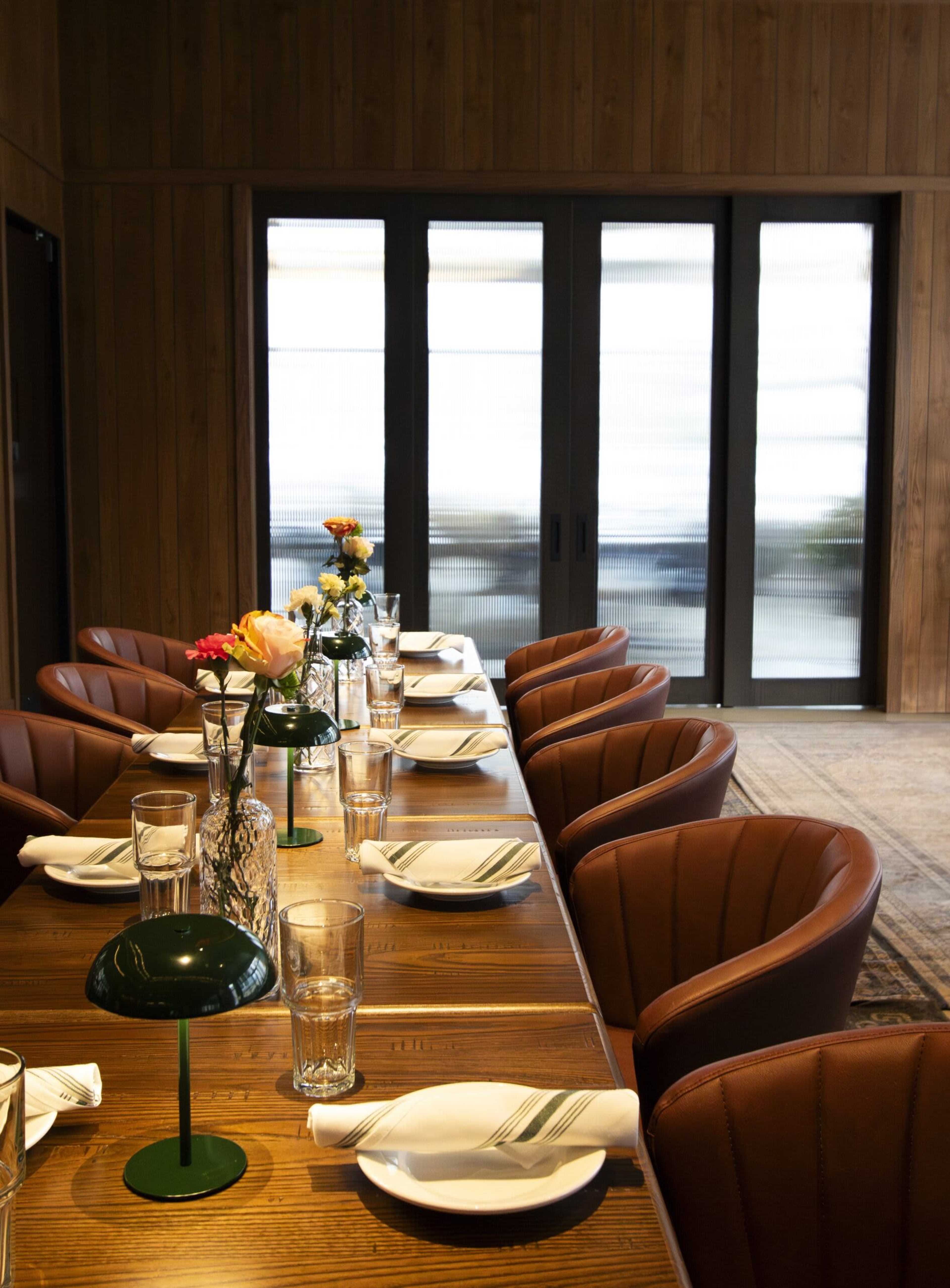 A long wooden dining table is set with plates, glasses, and floral arrangements, surrounded by brown leather chairs in a warmly decorated room with large windows.