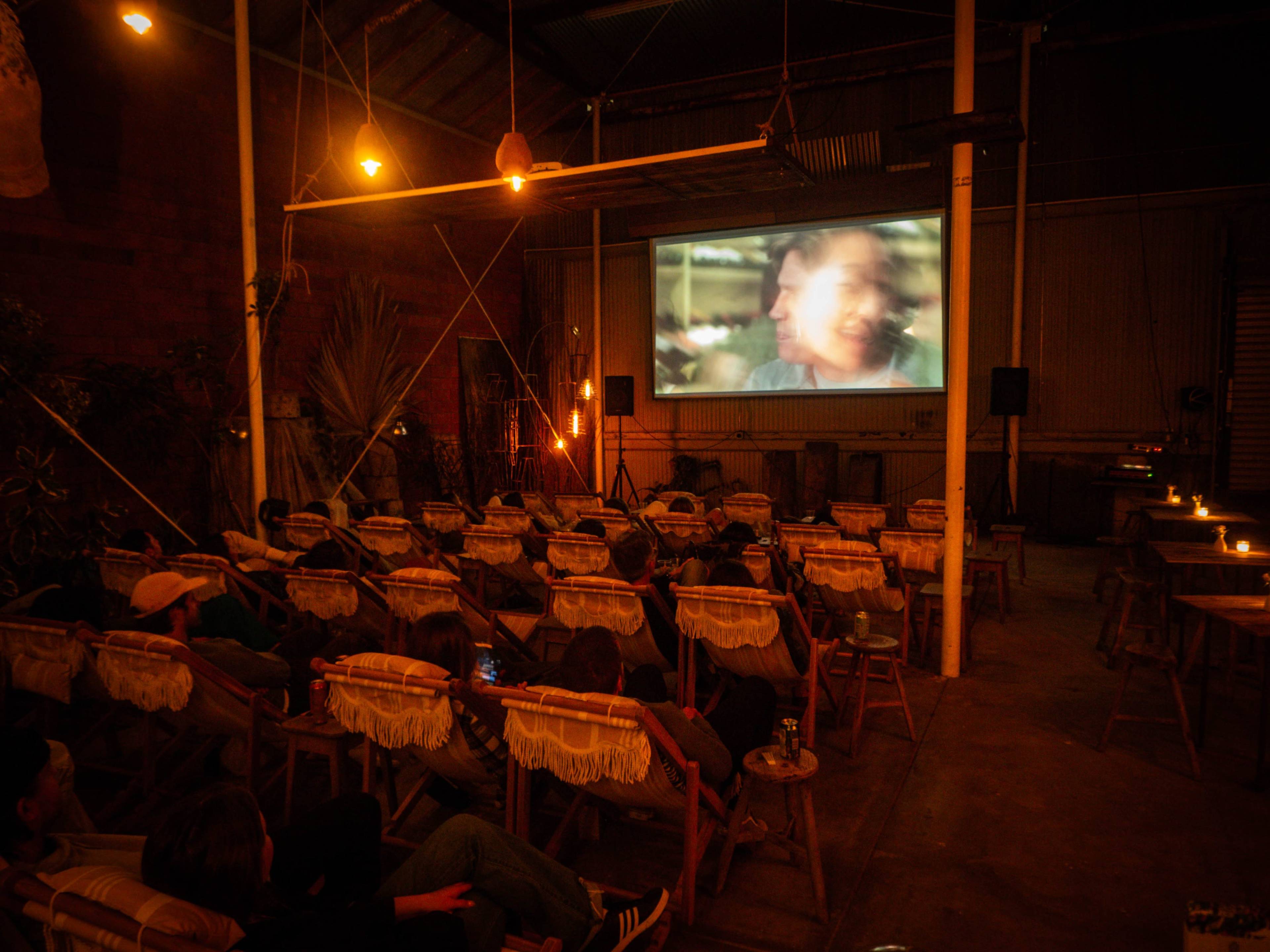 A dimly lit indoor space features a screen showing a film, with rows of wooden seating occupied by viewers.
