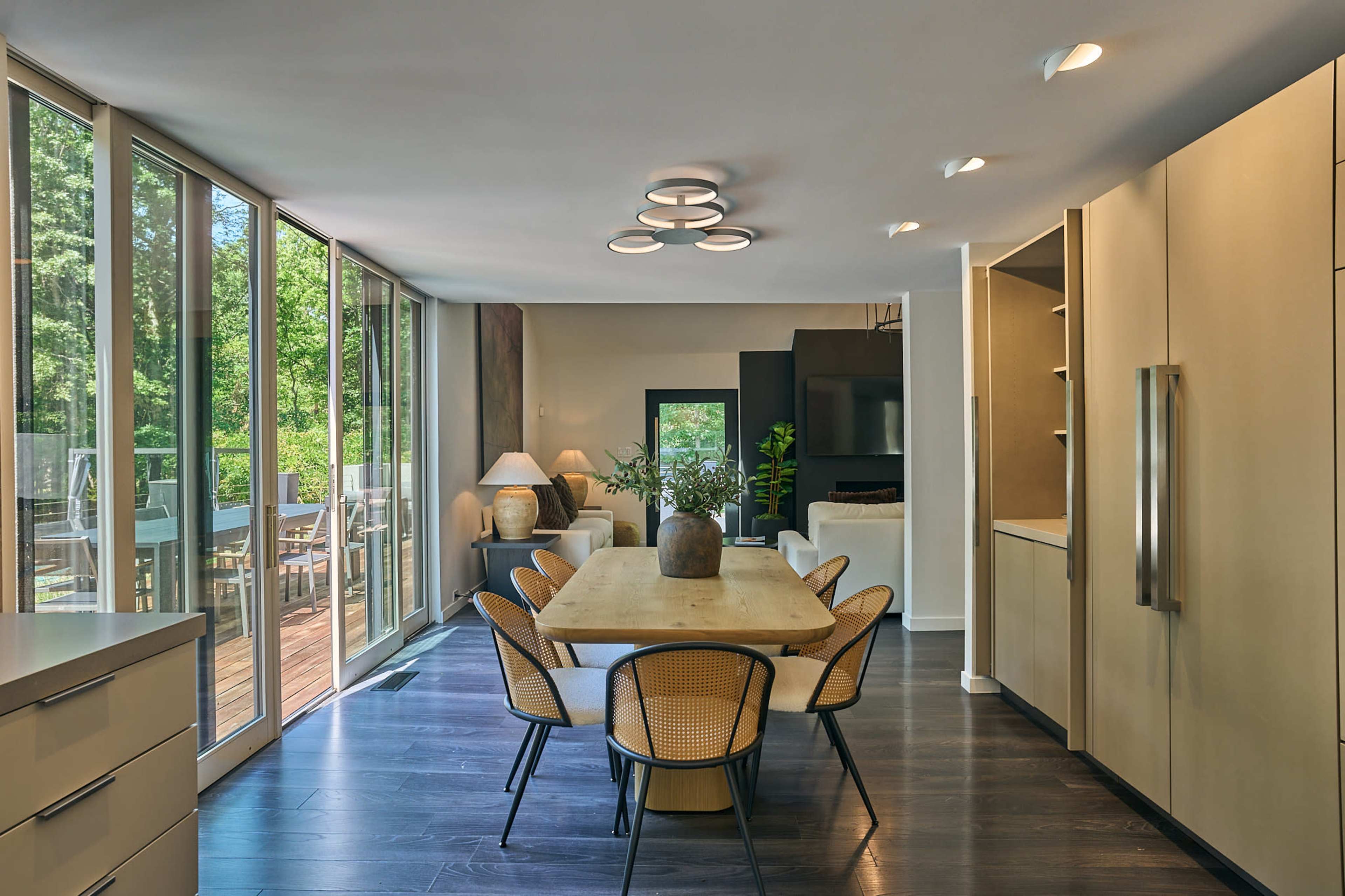 The image shows a modern dining area with a wooden table surrounded by six chairs, large windows letting in natural light, and a view of an outdoor deck.