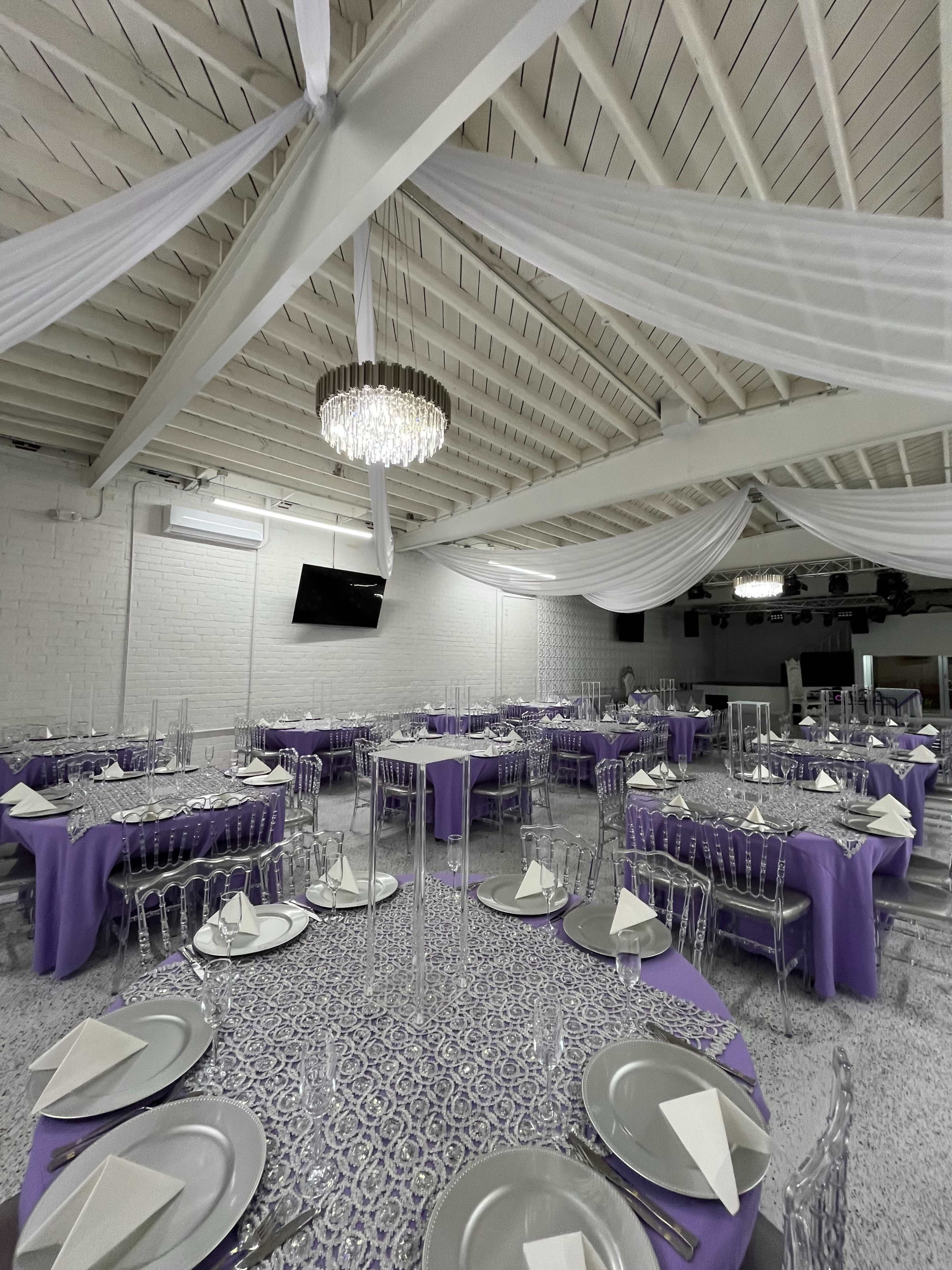 The image shows a spacious event room set up for a banquet, featuring tables dressed in purple tablecloths, elegant place settings, and a chandelier hanging from the ceiling.