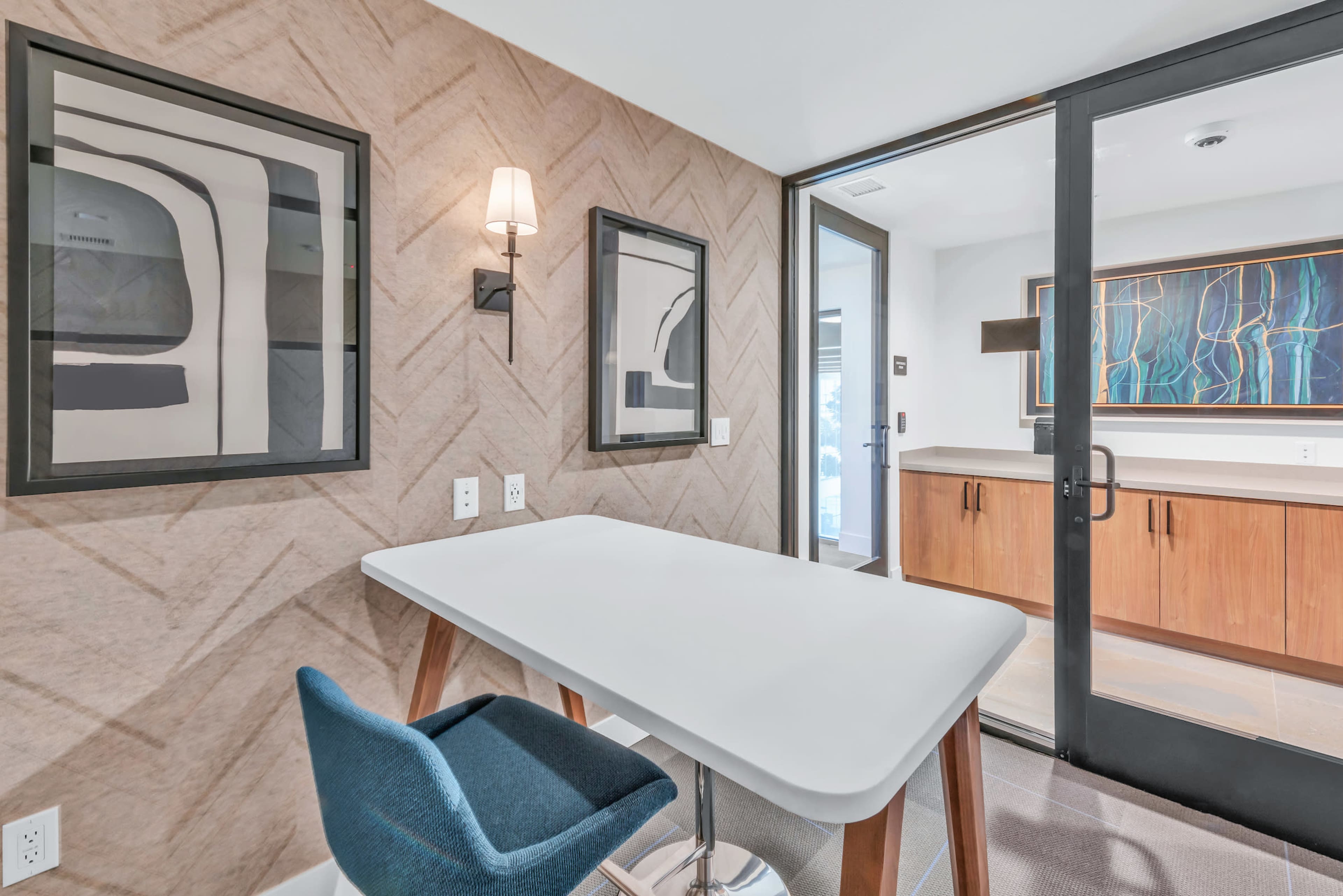A modern office space features a minimalist white table, a blue chair, framed abstract art on the walls, and a glass door leading to an adjacent area.