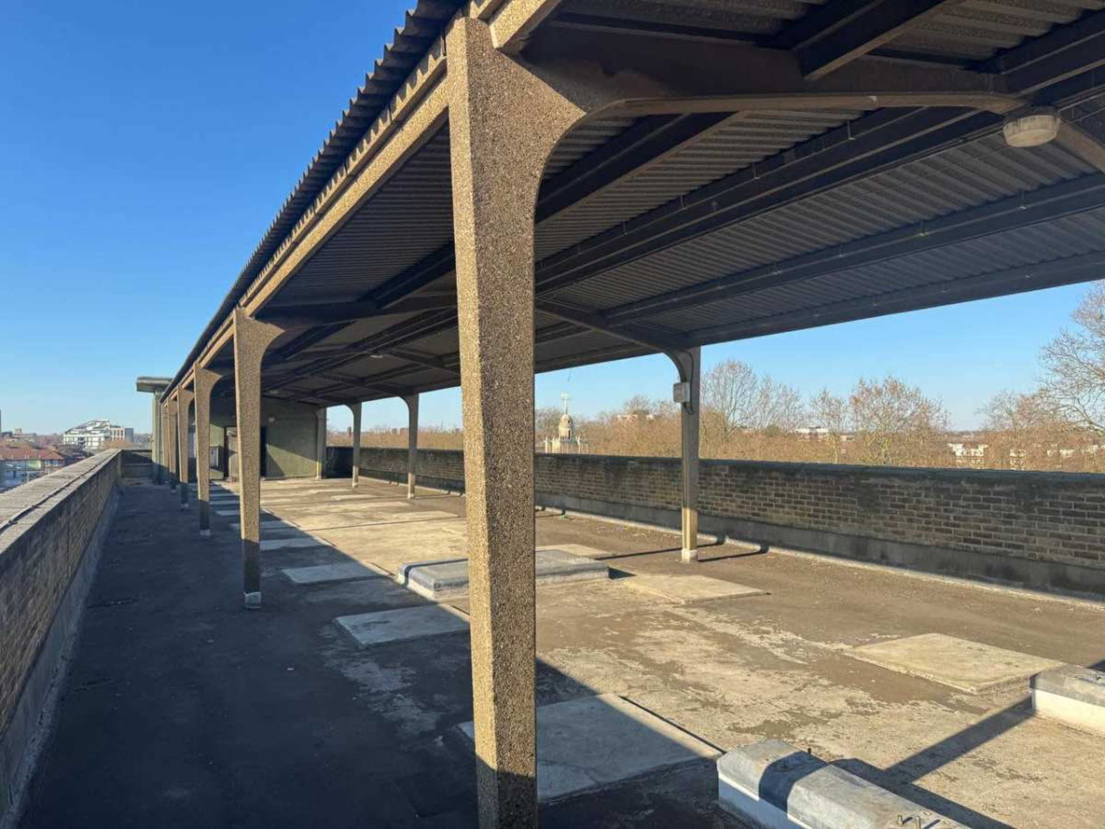 The image shows a vacant, covered parking area with concrete support beams and a clear blue sky overhead.
