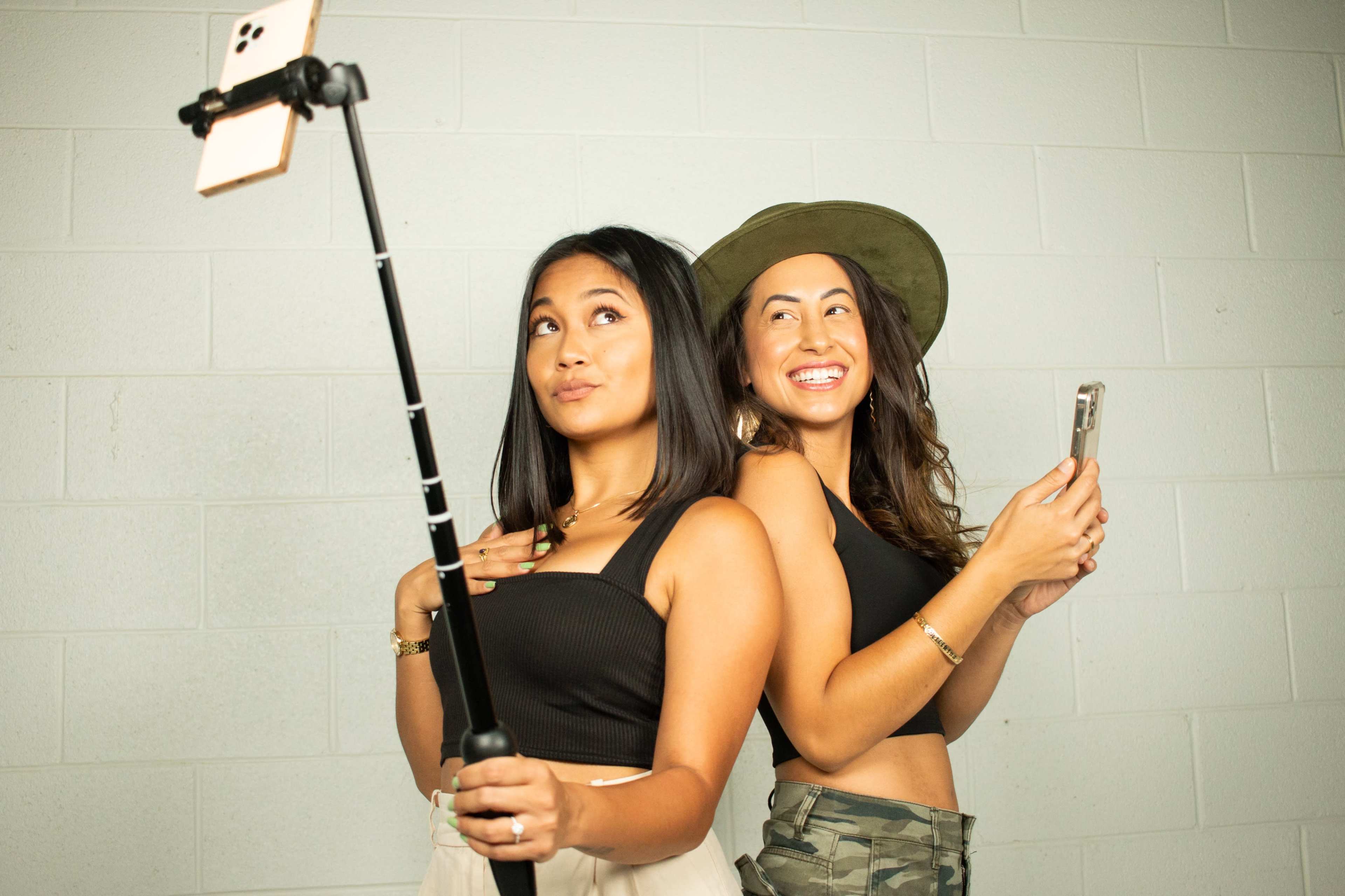 Two women pose for a selfie with a smartphone on a stick, smiling and facing different directions against a plain gray wall.