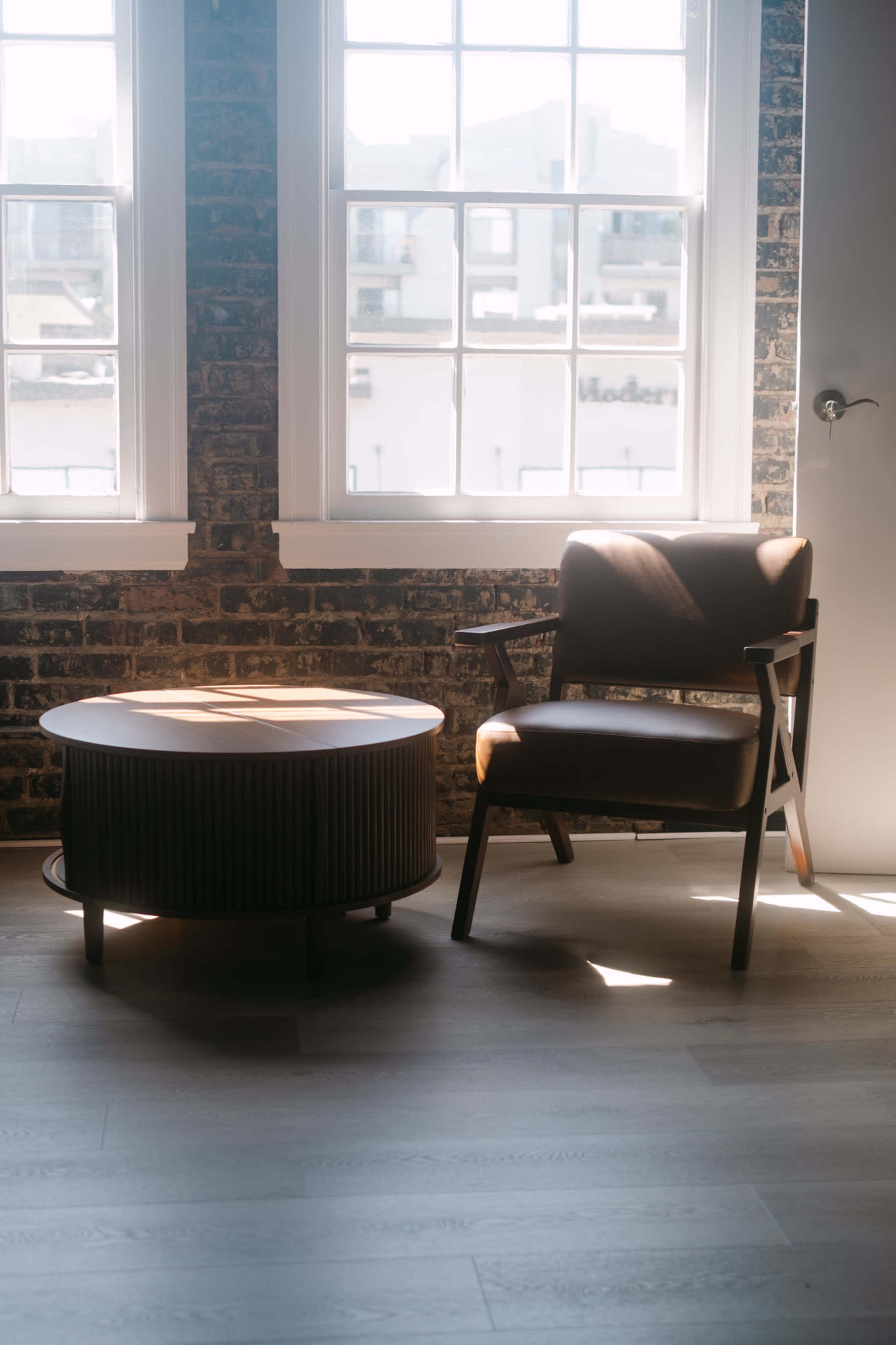 A wooden chair and round coffee table are positioned beside large windows in a room with exposed brick walls.