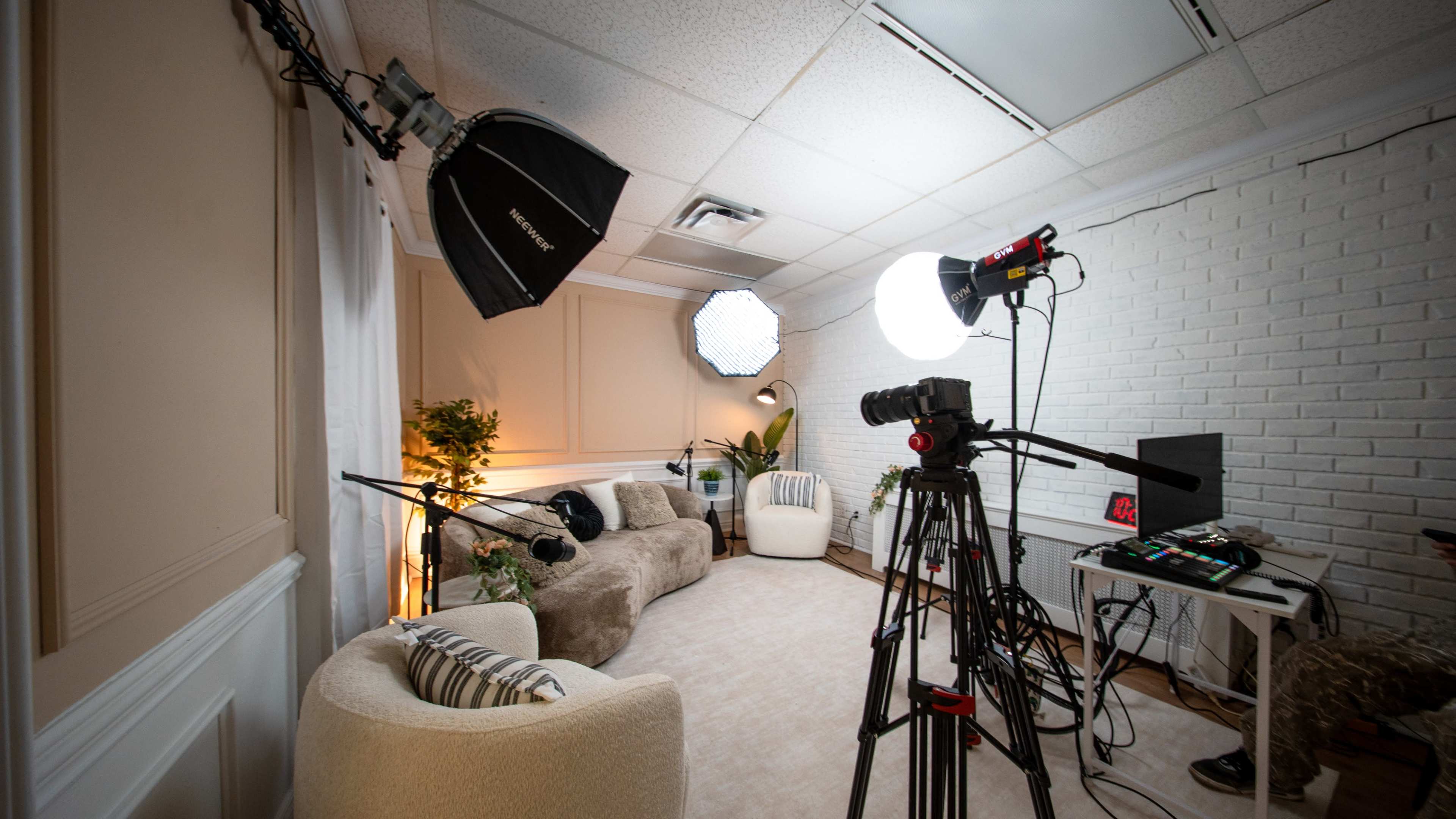 The image shows a well-lit studio setup featuring two softbox lights, a video camera on a tripod, and a cozy seating arrangement against a textured white wall.