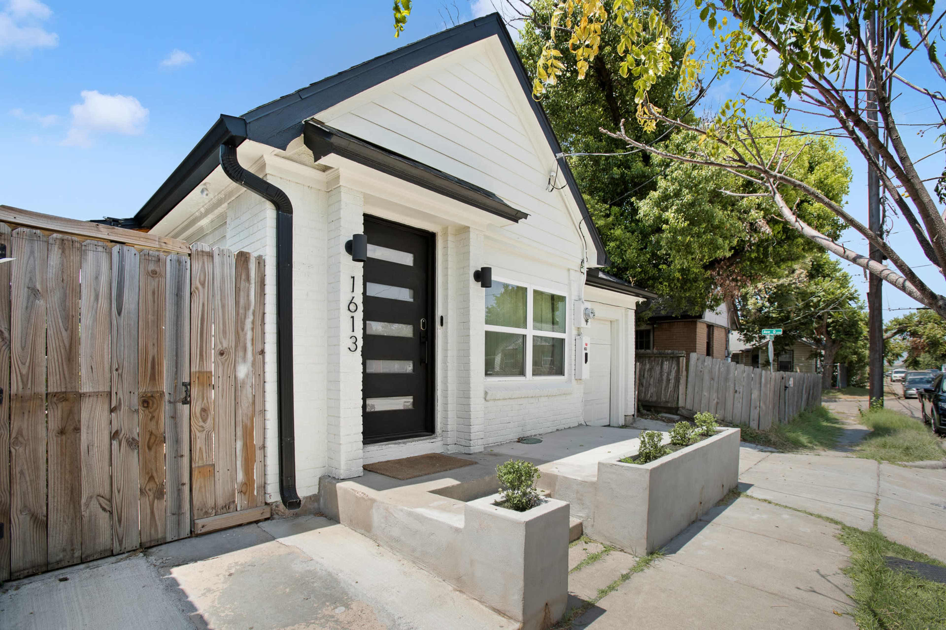The image shows a white, single-story house with a black door, a fenced yard, and a small concrete staircase leading to the entrance.