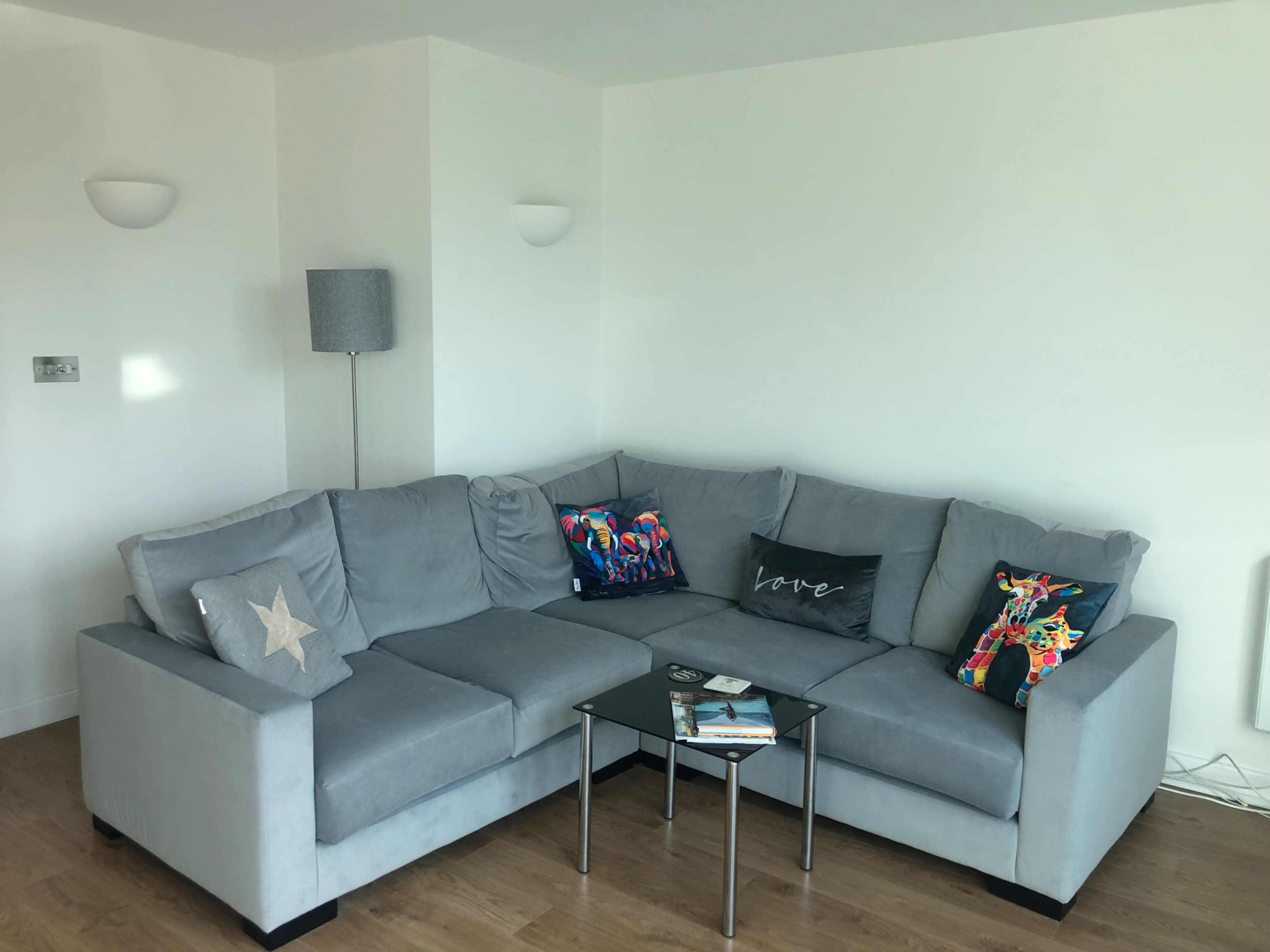 A light gray sectional sofa with decorative pillows, accompanied by a glass coffee table on a wooden floor in a bright room with white walls.