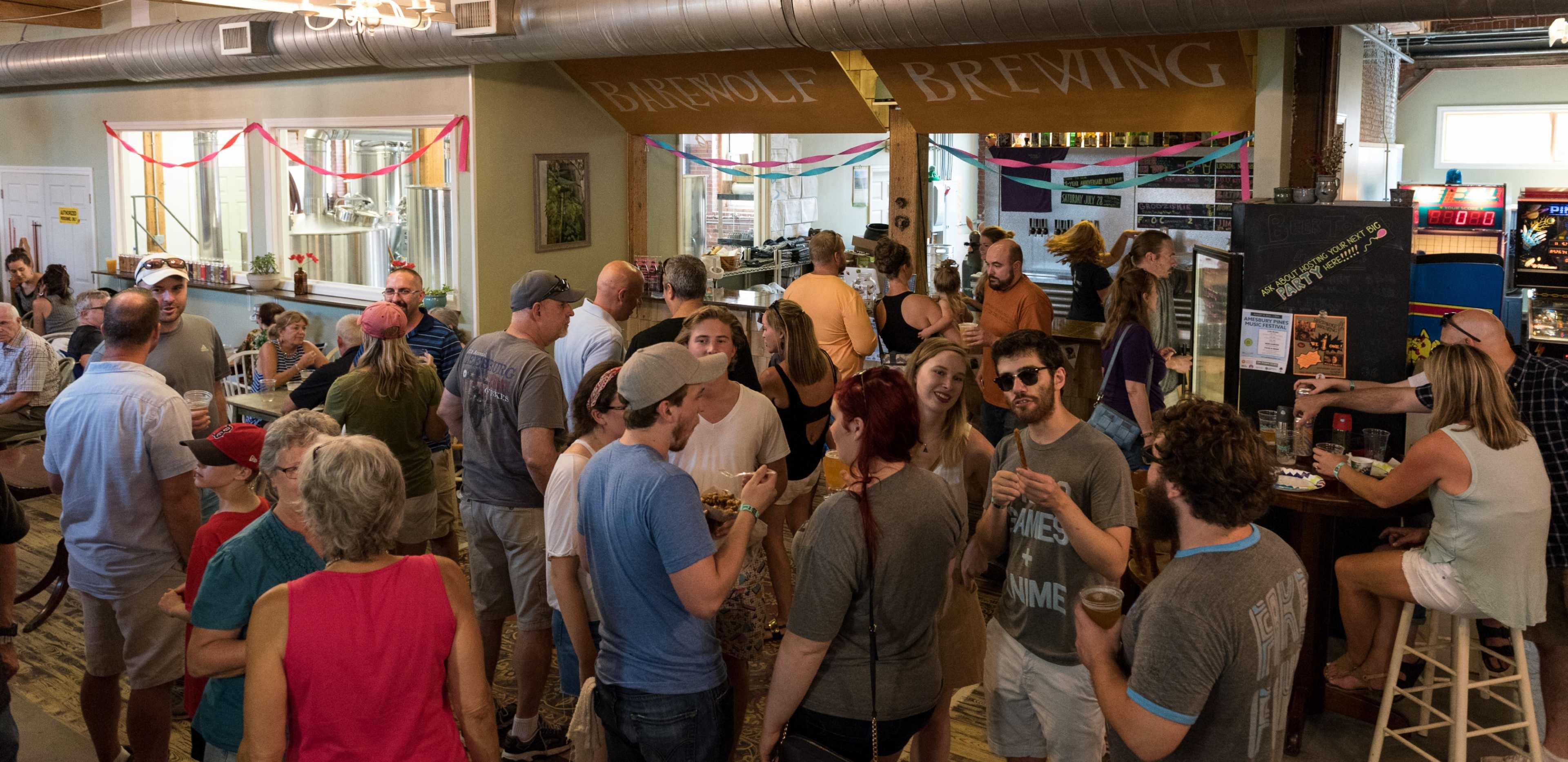 A crowd of people socializes inside a brewery, with some enjoying drinks and food at tables while others gather in clusters.