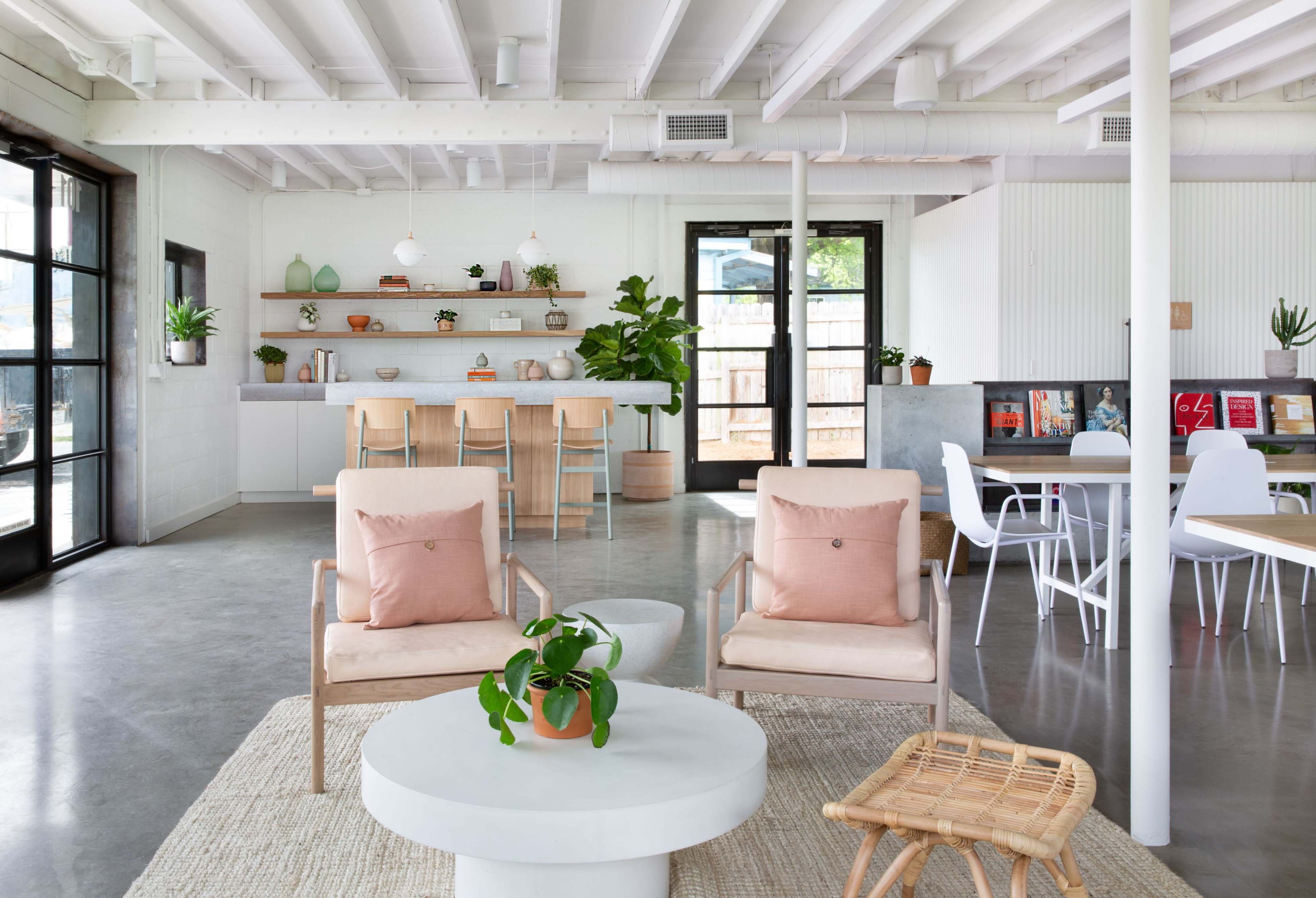 The image shows a modern, open-concept living space featuring light-colored walls, a kitchen area with bar seating, and various plants, along with seating arrangements including comfortable chairs and a round coffee table.