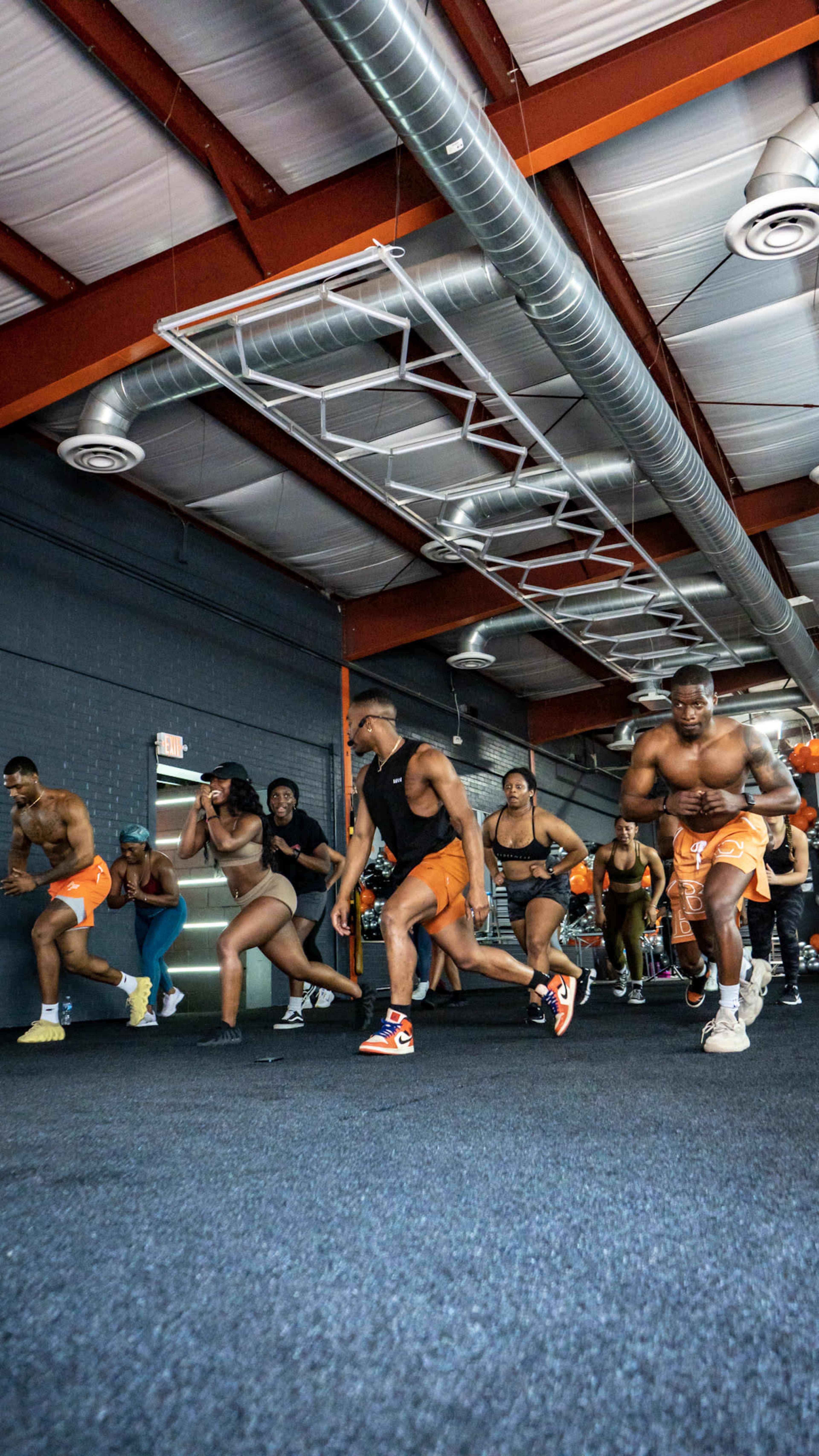 A group of people participates in a high-energy workout session inside a gym with exposed metal beams and fitness equipment in the background.