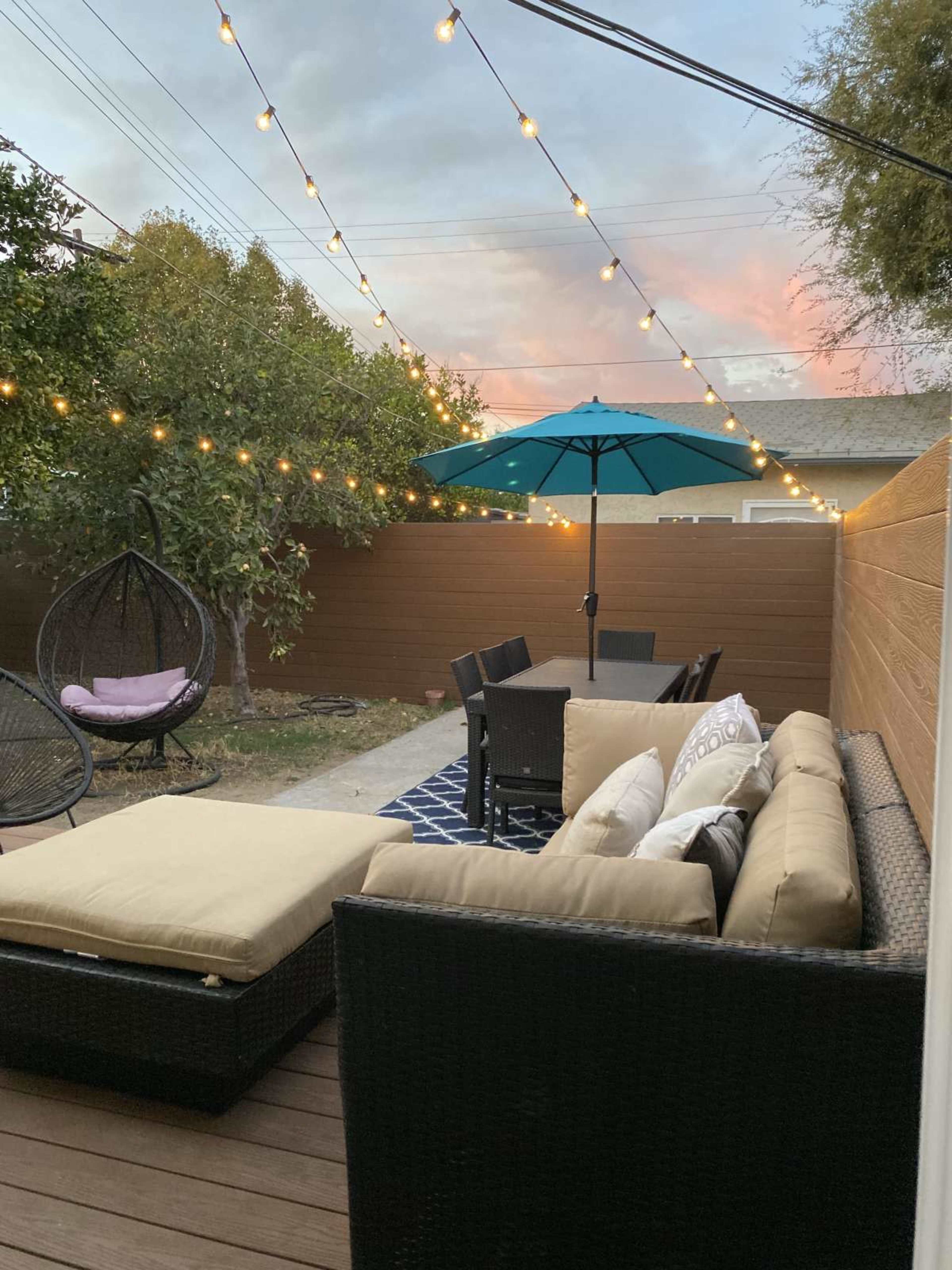 A cozy backyard patio features a sectional sofa, a dining table with chairs, and an umbrella, all illuminated by string lights overhead.