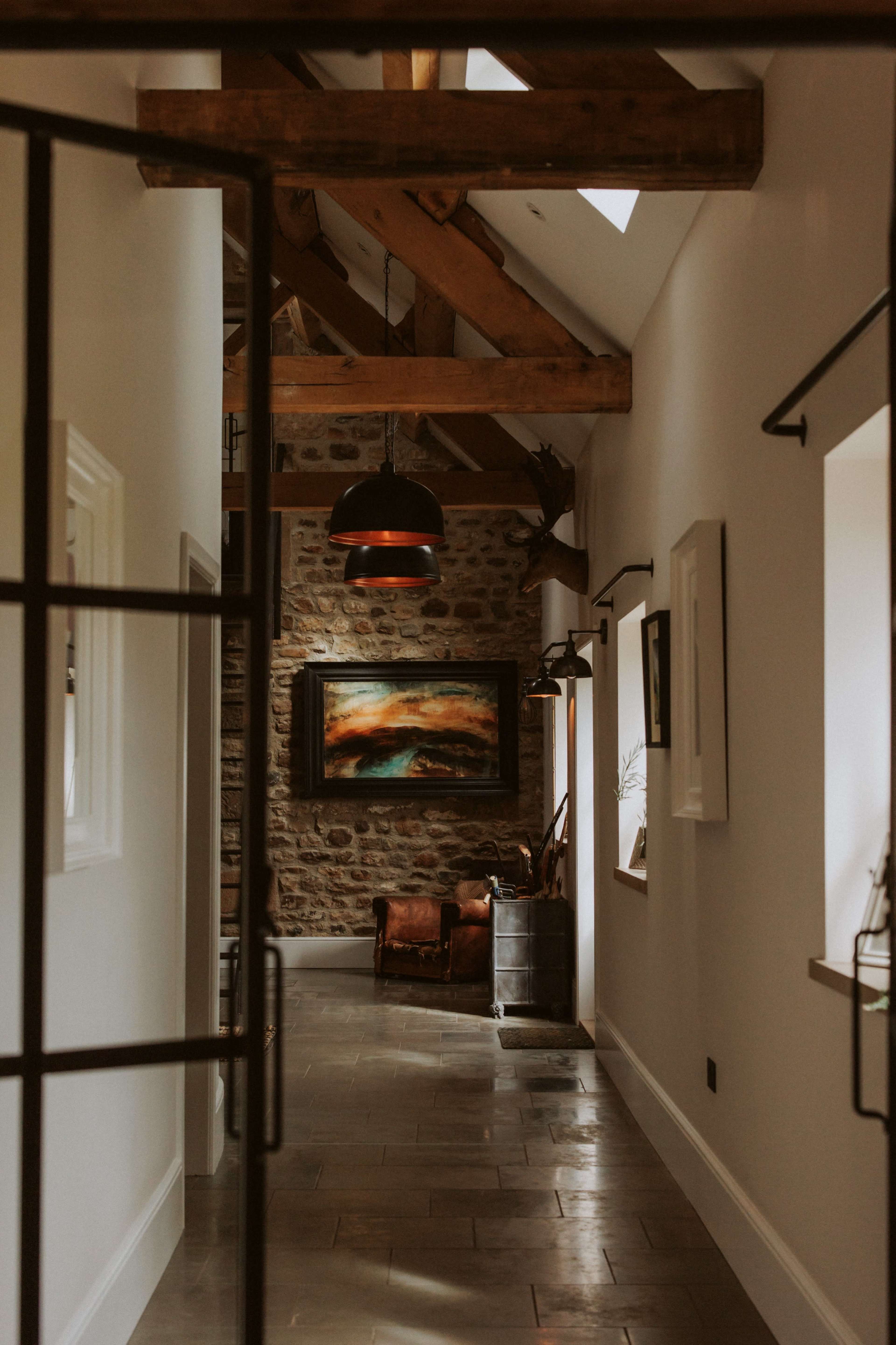 A hallway featuring exposed wooden beams, stone walls, and a large painting at the end, with a leather armchair and industrial light fixtures.