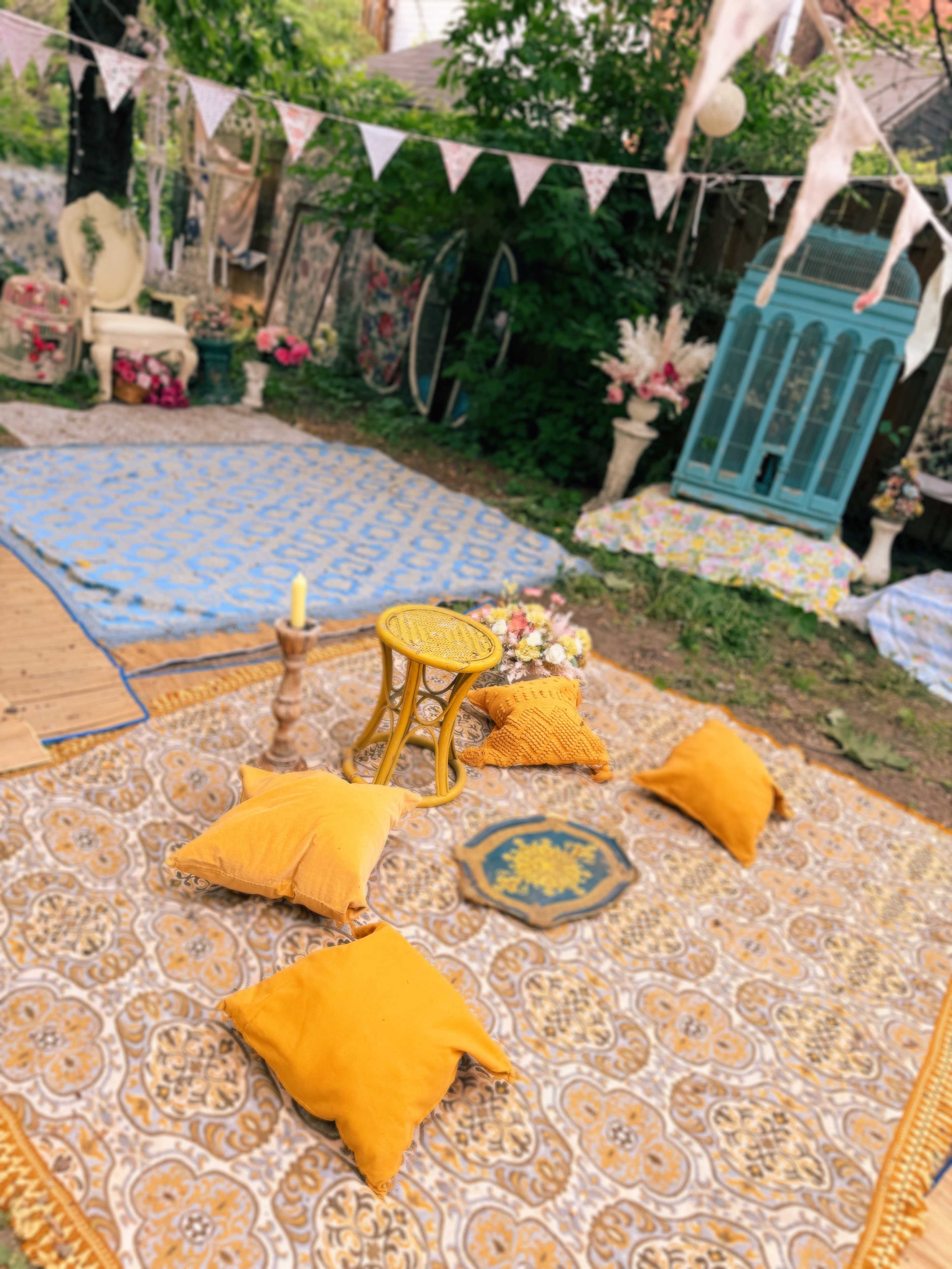 The image shows a cozy outdoor space with patterned rugs, yellow pillows, a small yellow table, and decorative items surrounded by greenery.