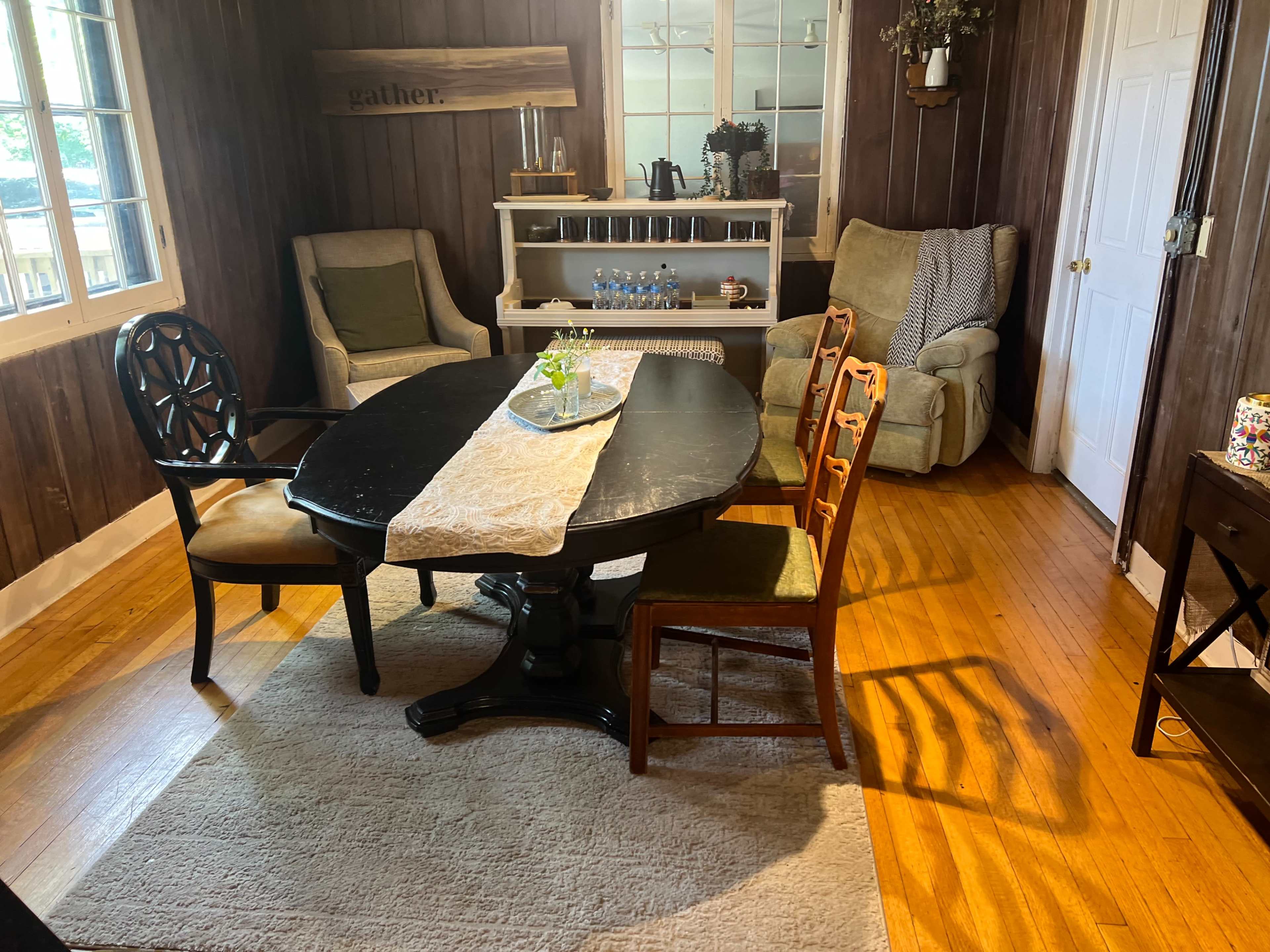 The image shows a dining area with a black round table, four chairs, a small side table, and a cabinet against a wooden wall.