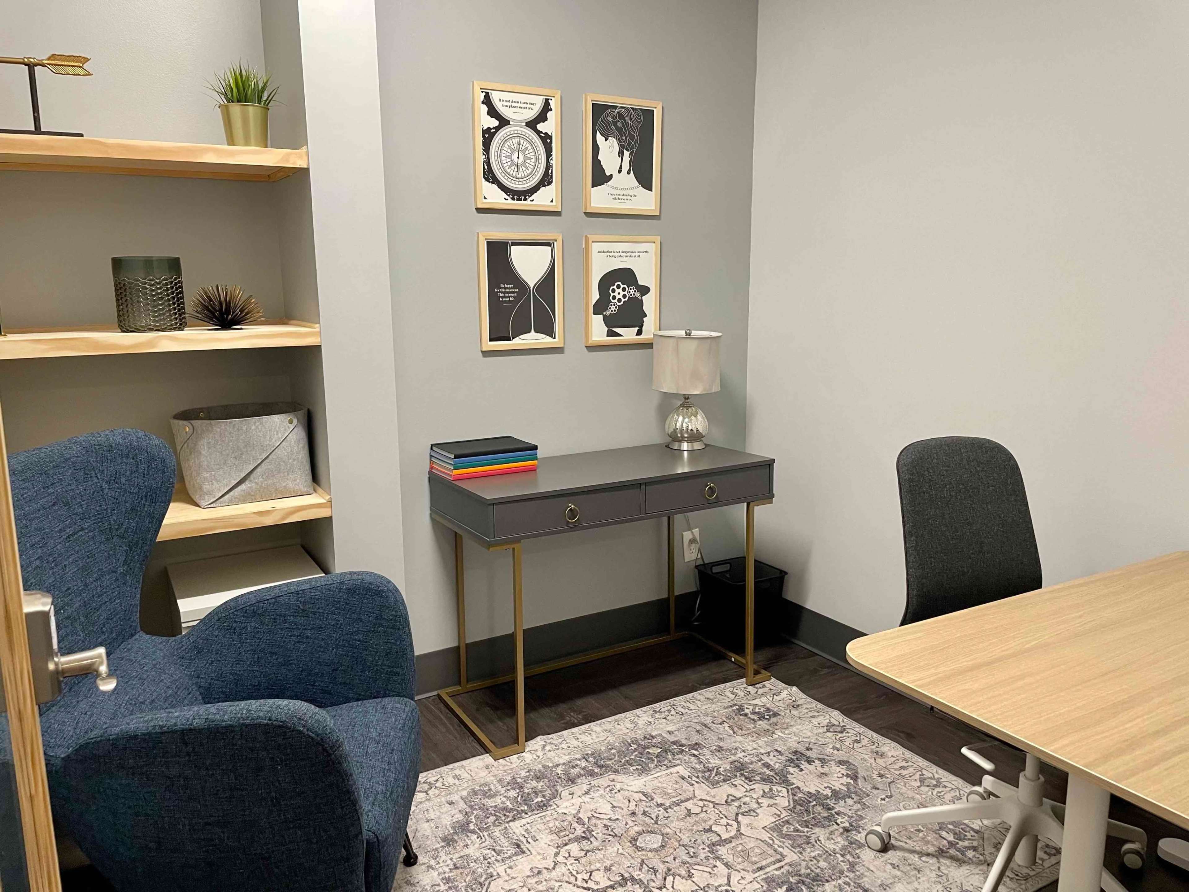 A small office space with a blue armchair, a gray desk with decorative items, and a wooden table, all set against a light gray wall.