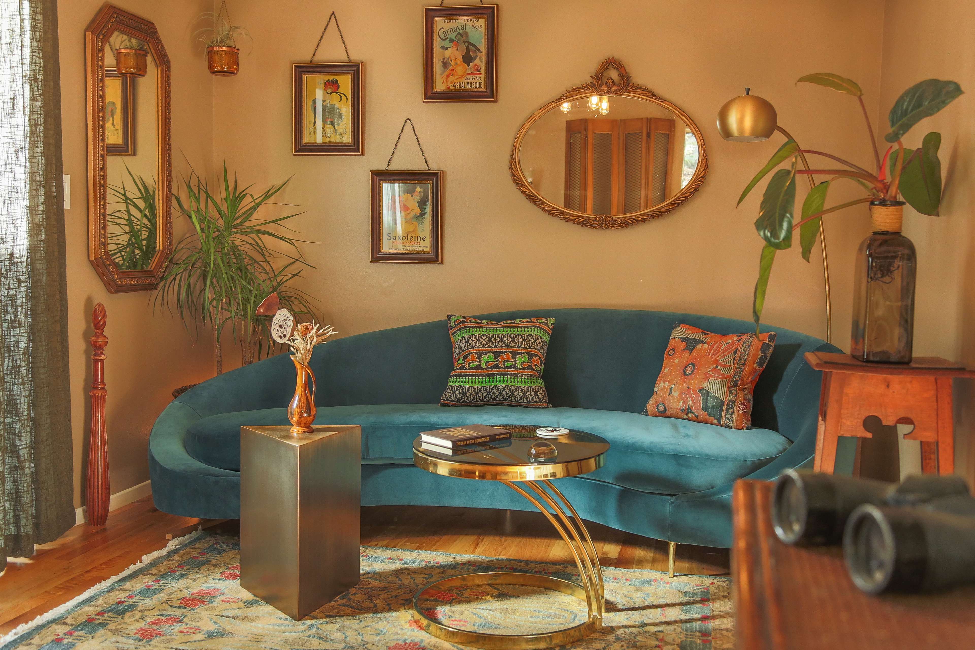 The scene features a curved teal sofa surrounded by framed artwork, a round coffee table, and decorative plants in a warmly lit living room.