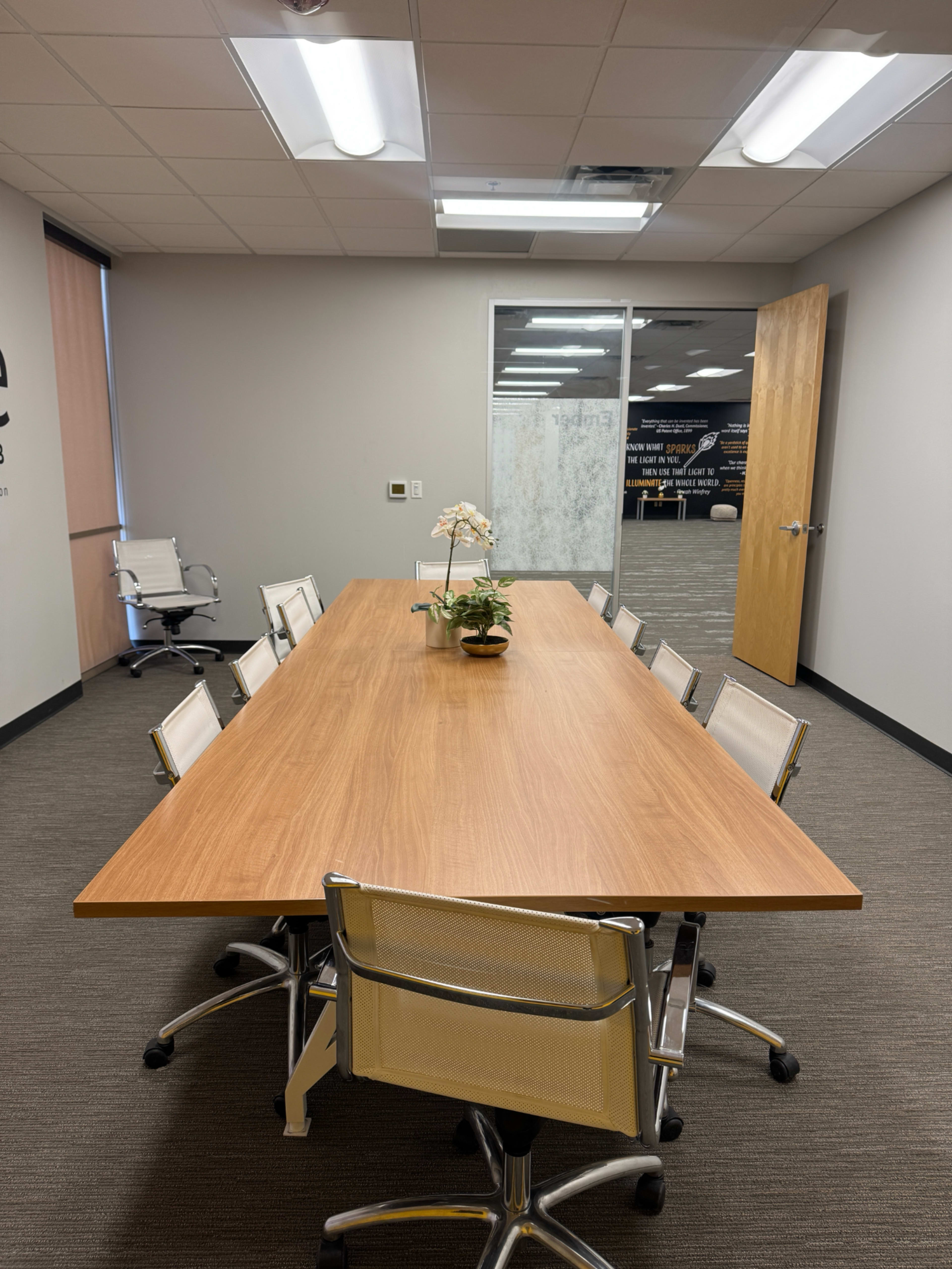 A large wooden conference table surrounded by eight white office chairs is set in a meeting room with gray walls and large windows.
