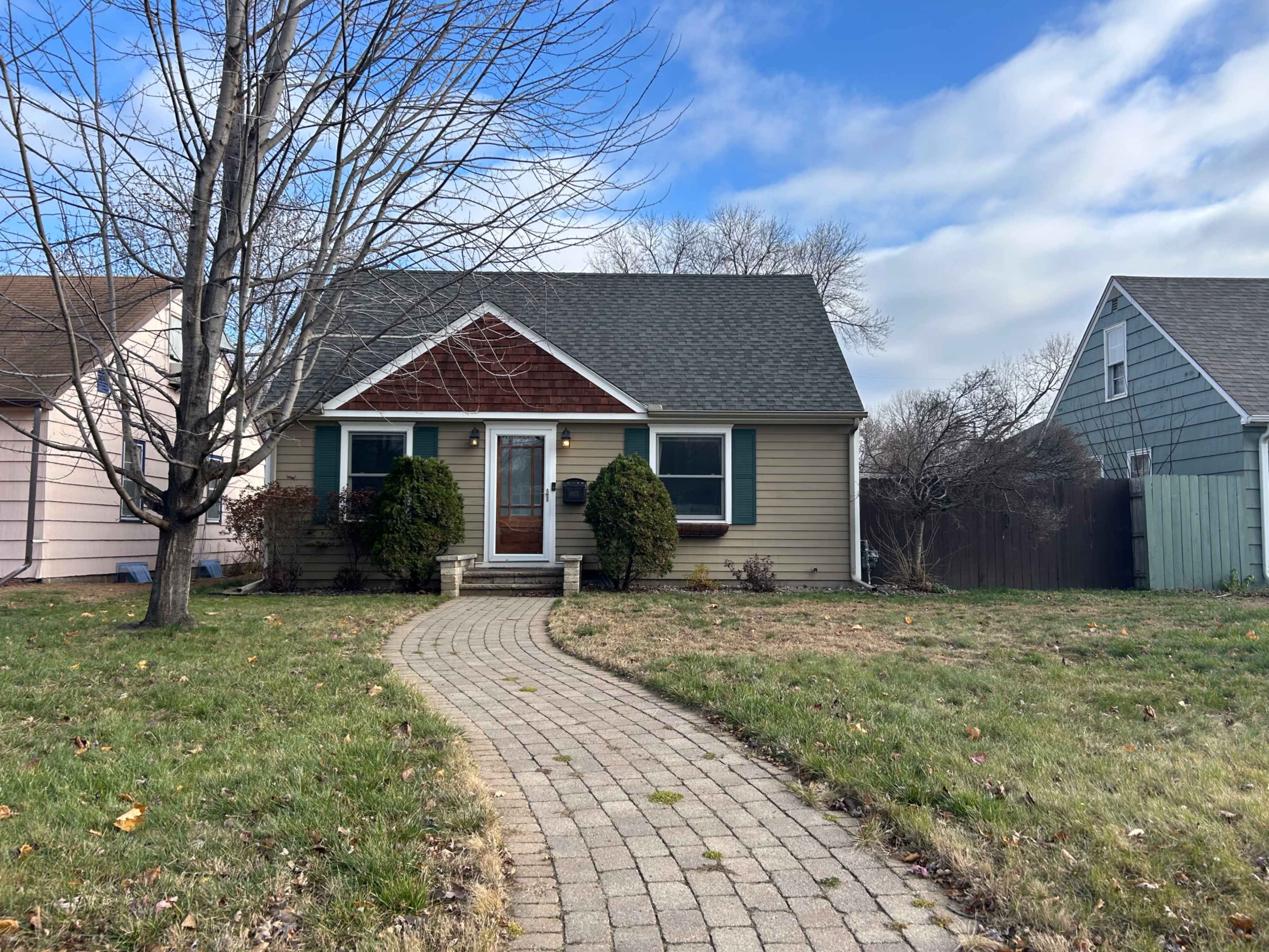The image shows a single-story house with a sloped roof, flanked by trees, and a brick pathway leading to the front door.