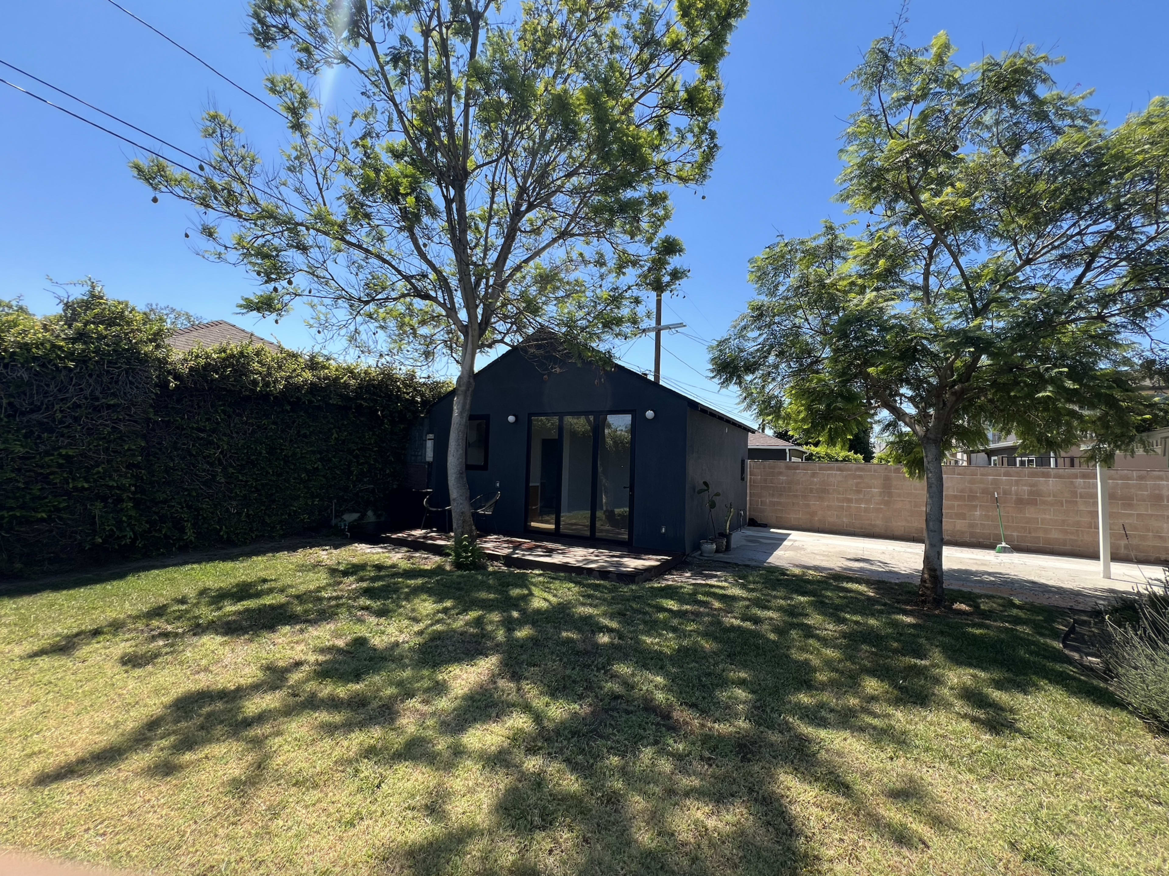 The image shows a small, dark-colored house with large windows, surrounded by a well-maintained lawn and trees, in a residential area under a clear sky.