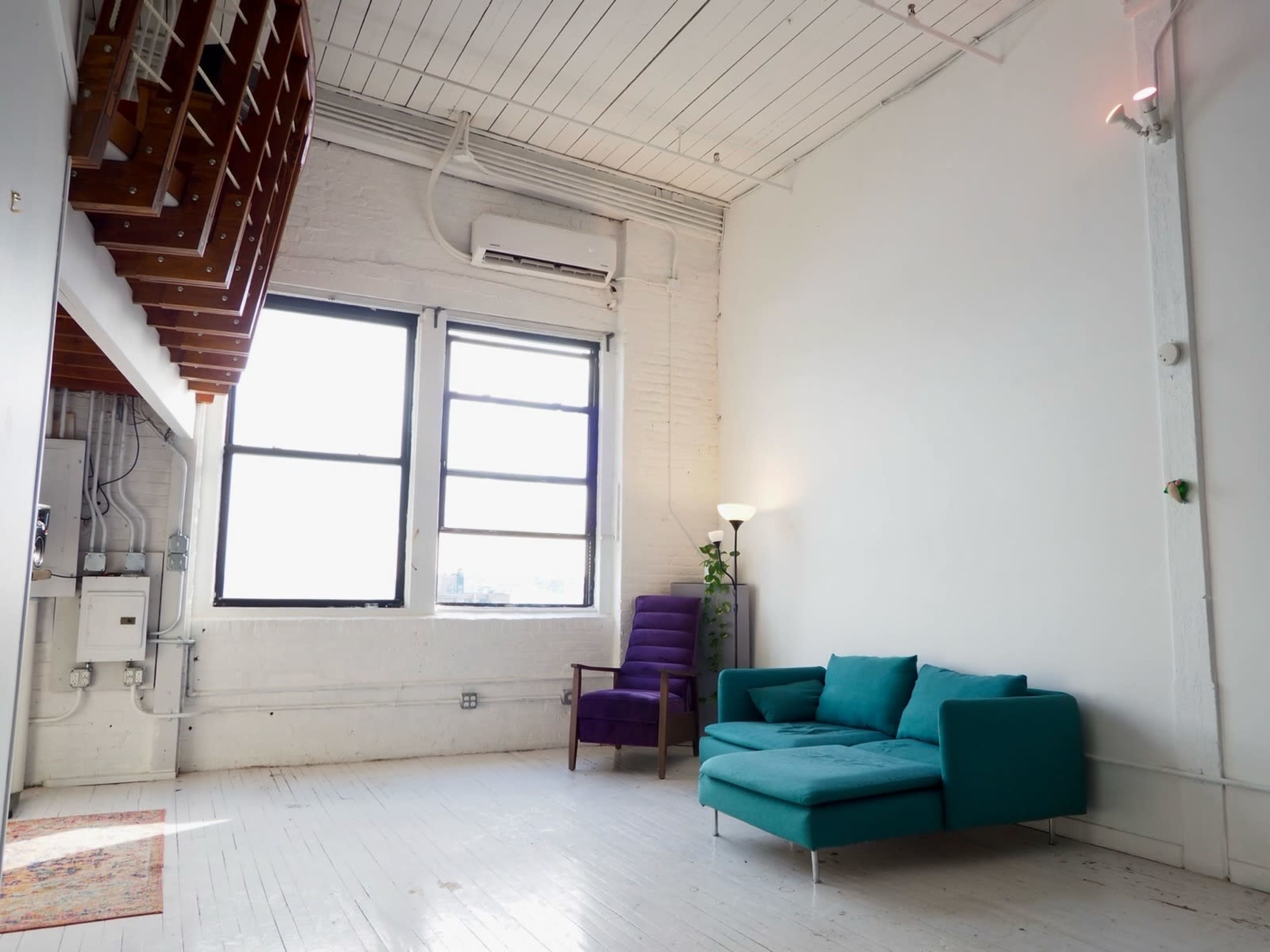 The image shows a bright, minimalist room featuring a blue sectional sofa, a purple armchair, and two large windows with white walls and wooden ceiling beams.