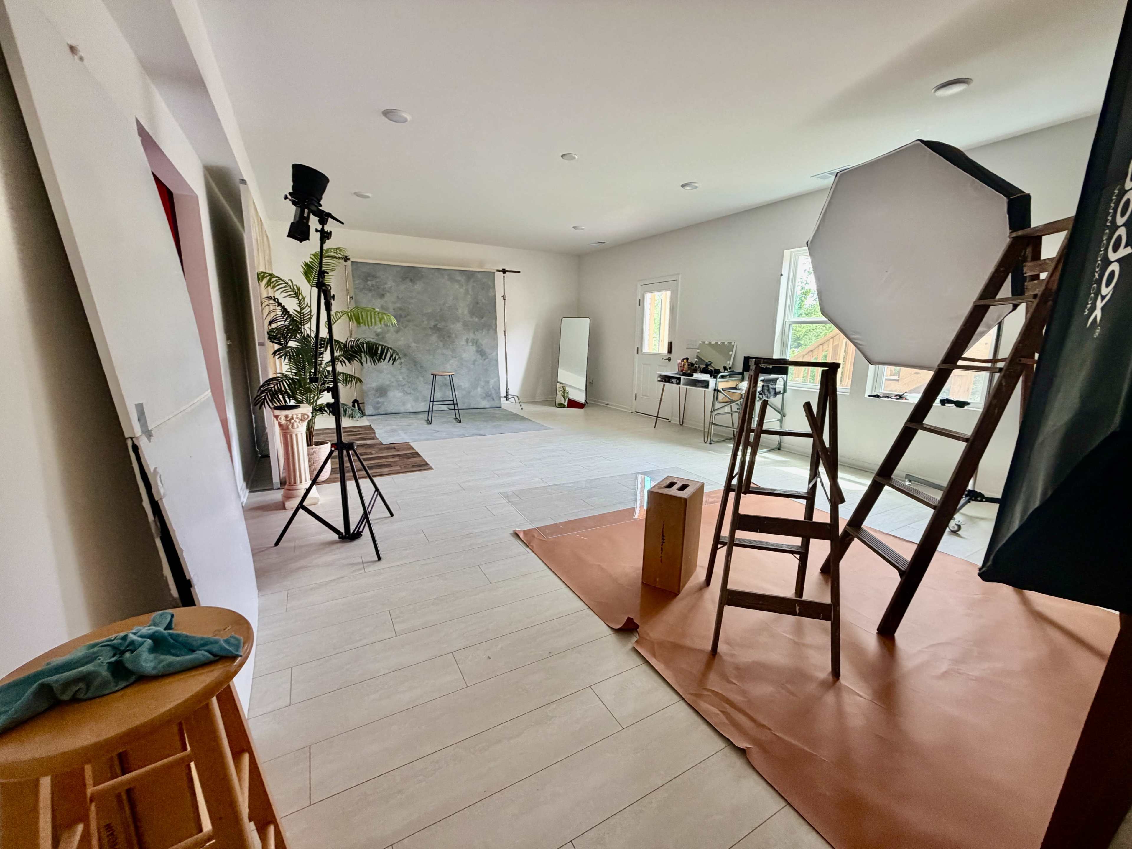 The image shows a spacious photography studio setting with various equipment, including a backdrop, lights, and props, arranged against white walls and wooden flooring.